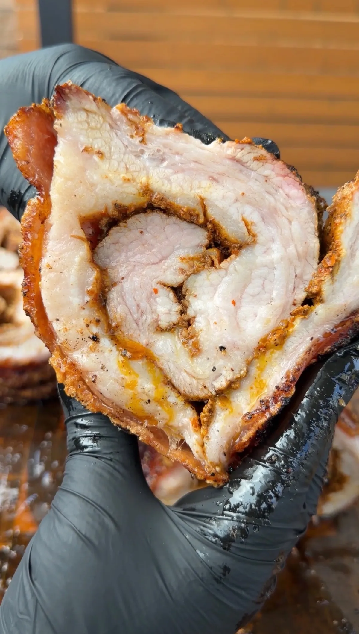 Hands holding a thick slice of smoked pork belly porchetta showing spiral layers of seasoned meat and crispy bark