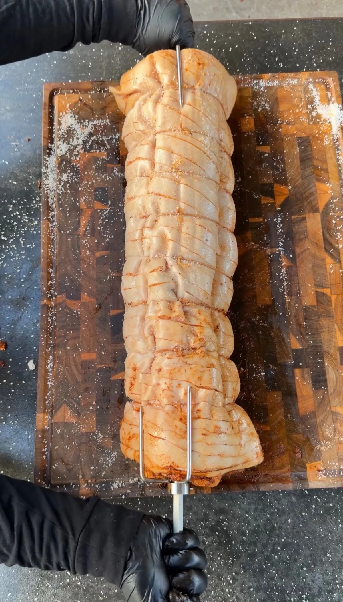 Scored pork belly porchetta on a rotisserie spit resting on a wooden cutting board, seasoned with salt
