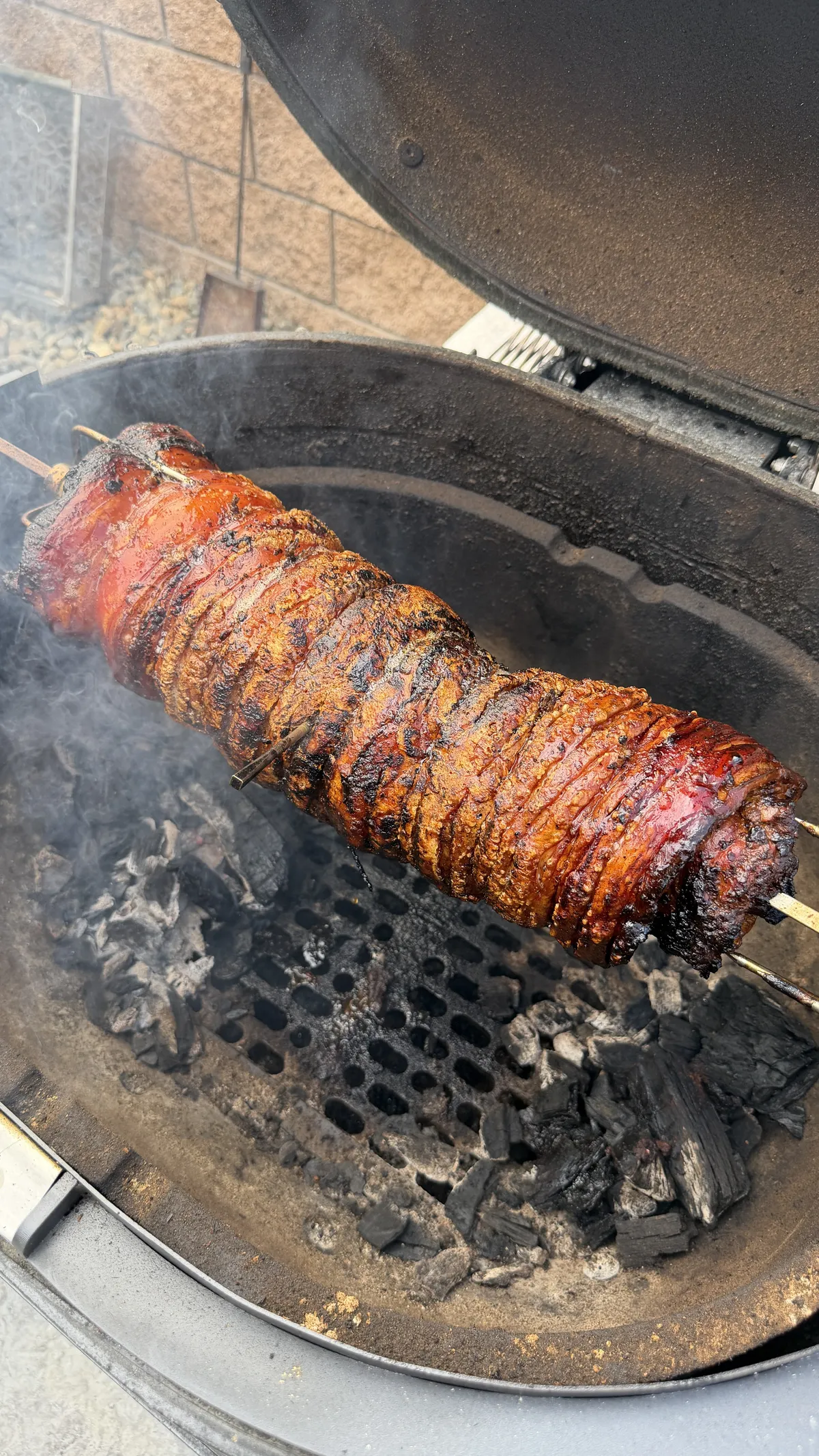 Rolled pork belly porchetta on skewers over charcoal in a kamado grill, with smoky caramelized crust