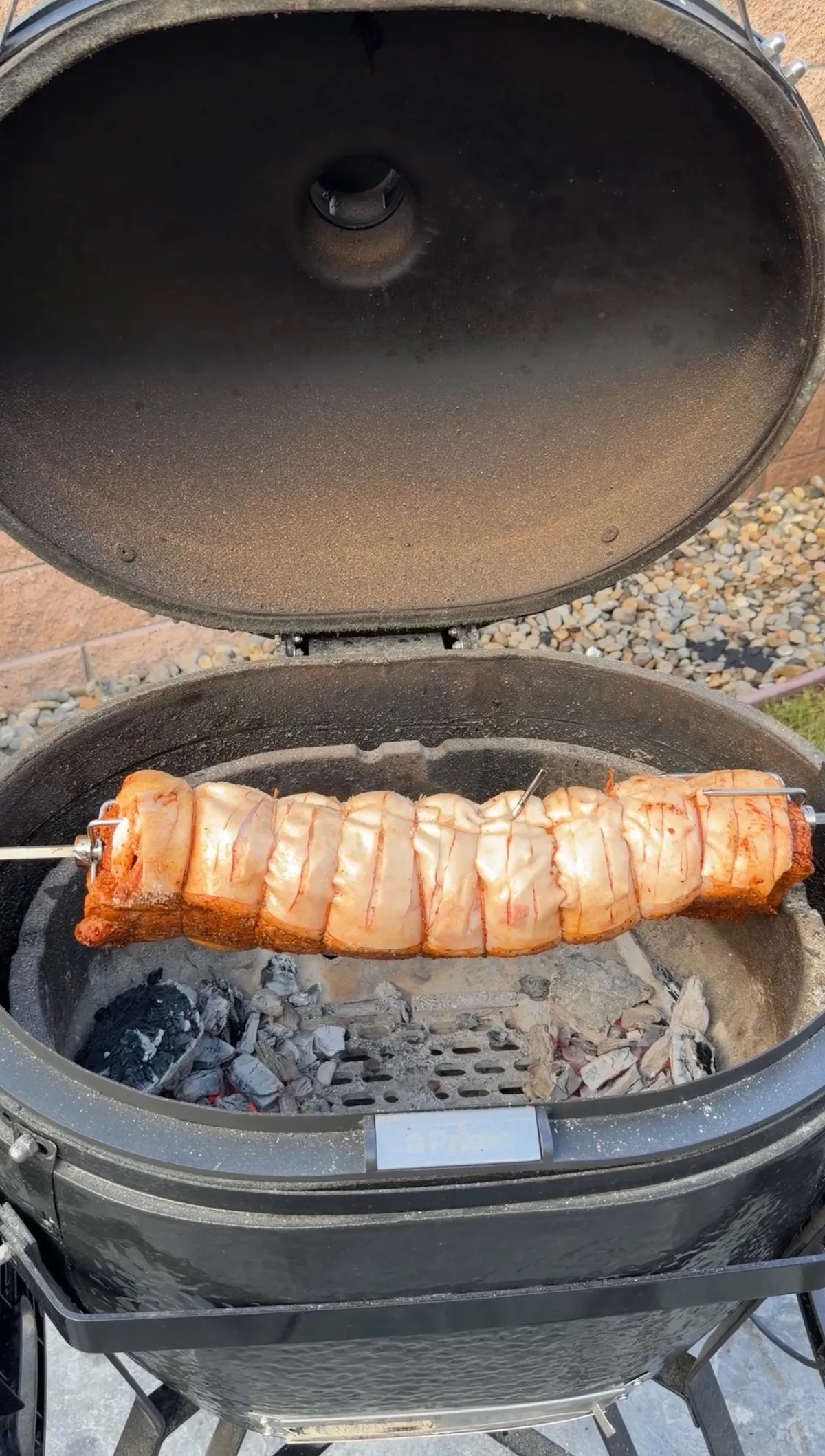 Rolled pork belly porchetta on a rotisserie spit cooking over charcoal in a kamado-style ceramic grill