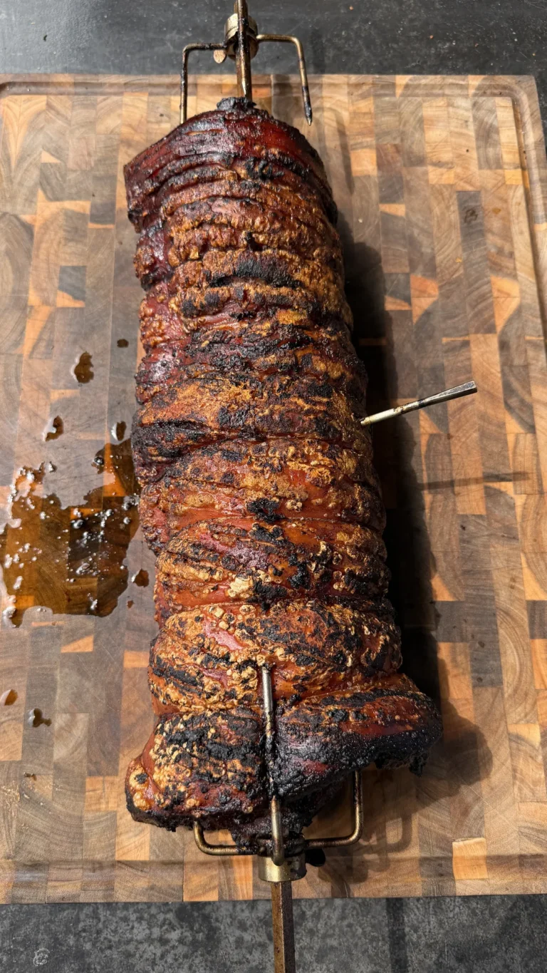 Whole pork belly porchetta on a rotisserie spit resting on a wooden cutting board with caramelized, charred crust