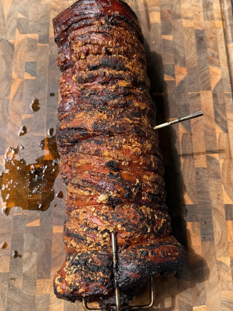 Whole pork belly porchetta on a rotisserie spit resting on a wooden cutting board with caramelized, charred crust