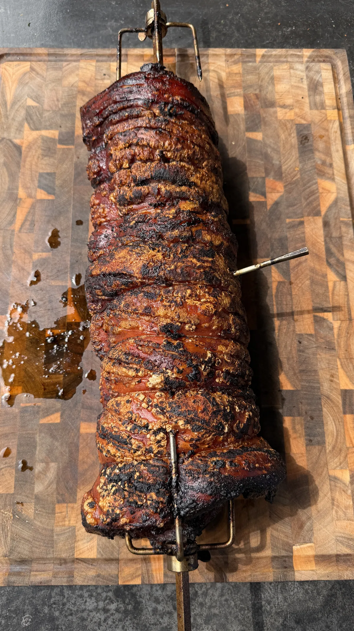Whole pork belly porchetta on a rotisserie spit resting on a wooden cutting board with caramelized, charred crust