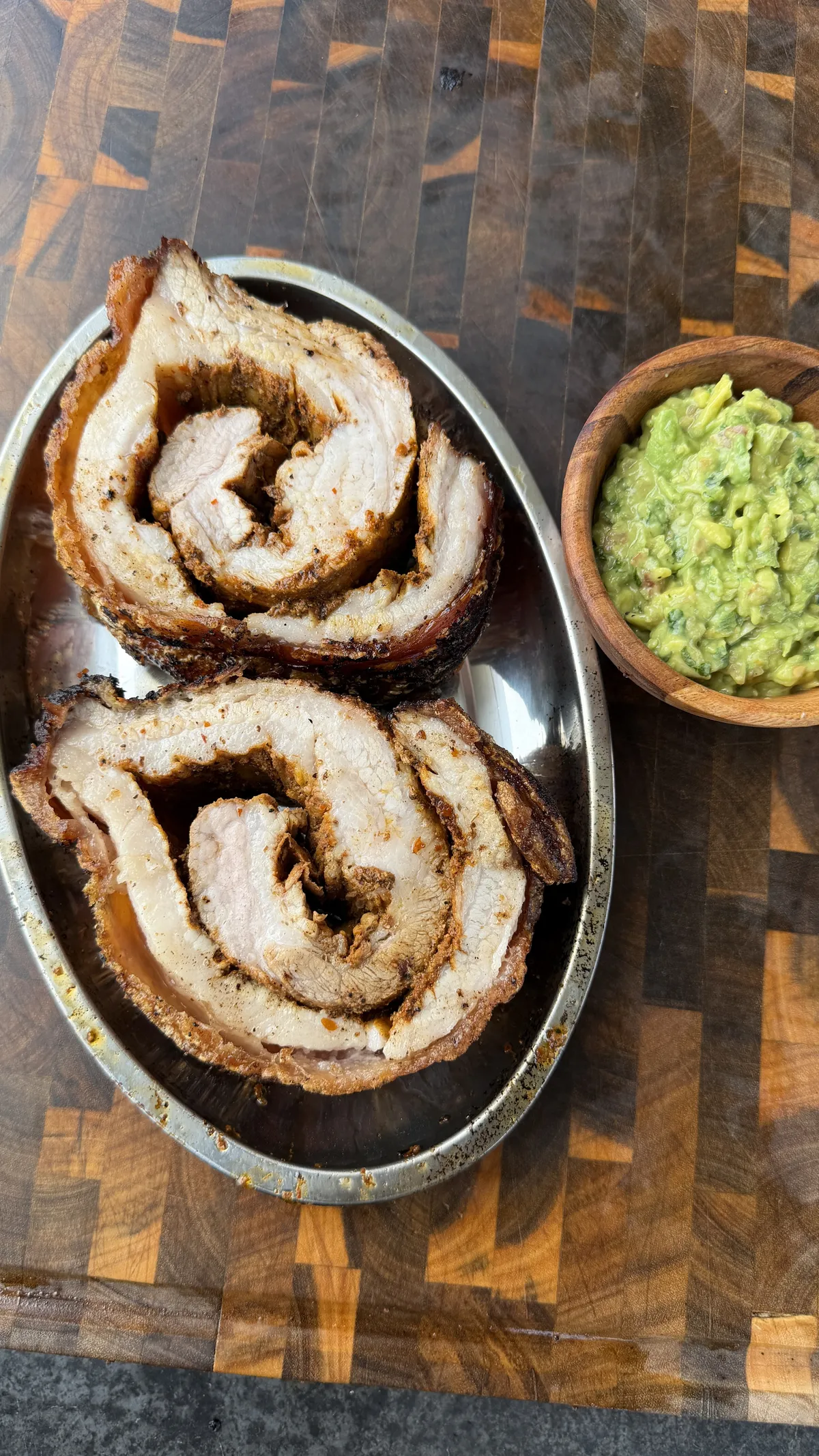 Sliced pork belly porchetta rolls on a silver platter served with a side of guacamole on a wooden board