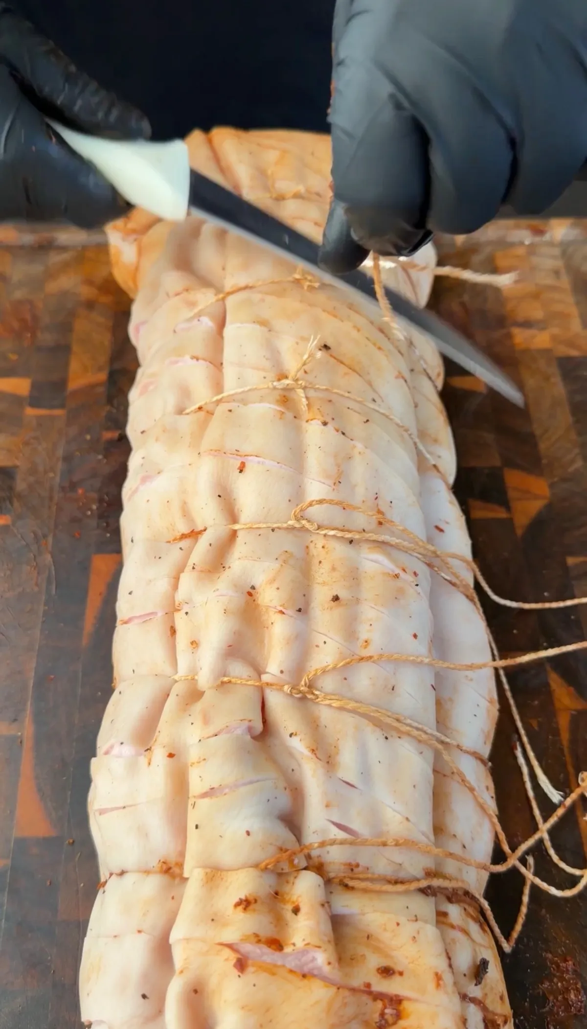Hands in black gloves cutting twine on a rolled pork belly porchetta scored and seasoned on a wooden board