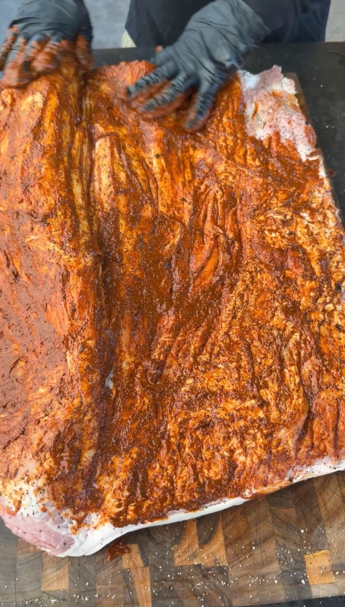 Hands in black gloves rubbing red spice paste onto a large flat pork belly on a wooden cutting board