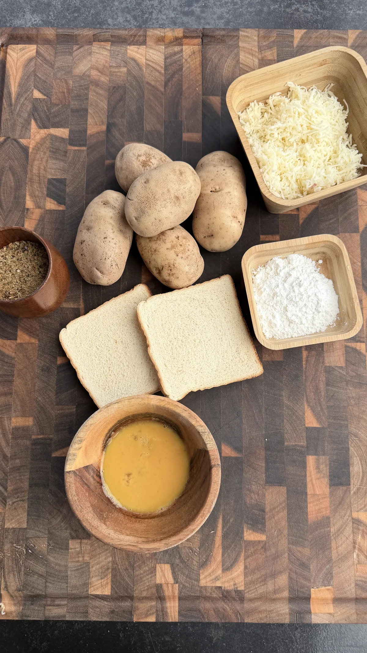 Ingredients for potato cheese balls: raw potatoes, shredded cheese, eggs, flour, bread slices, and spices on wooden board