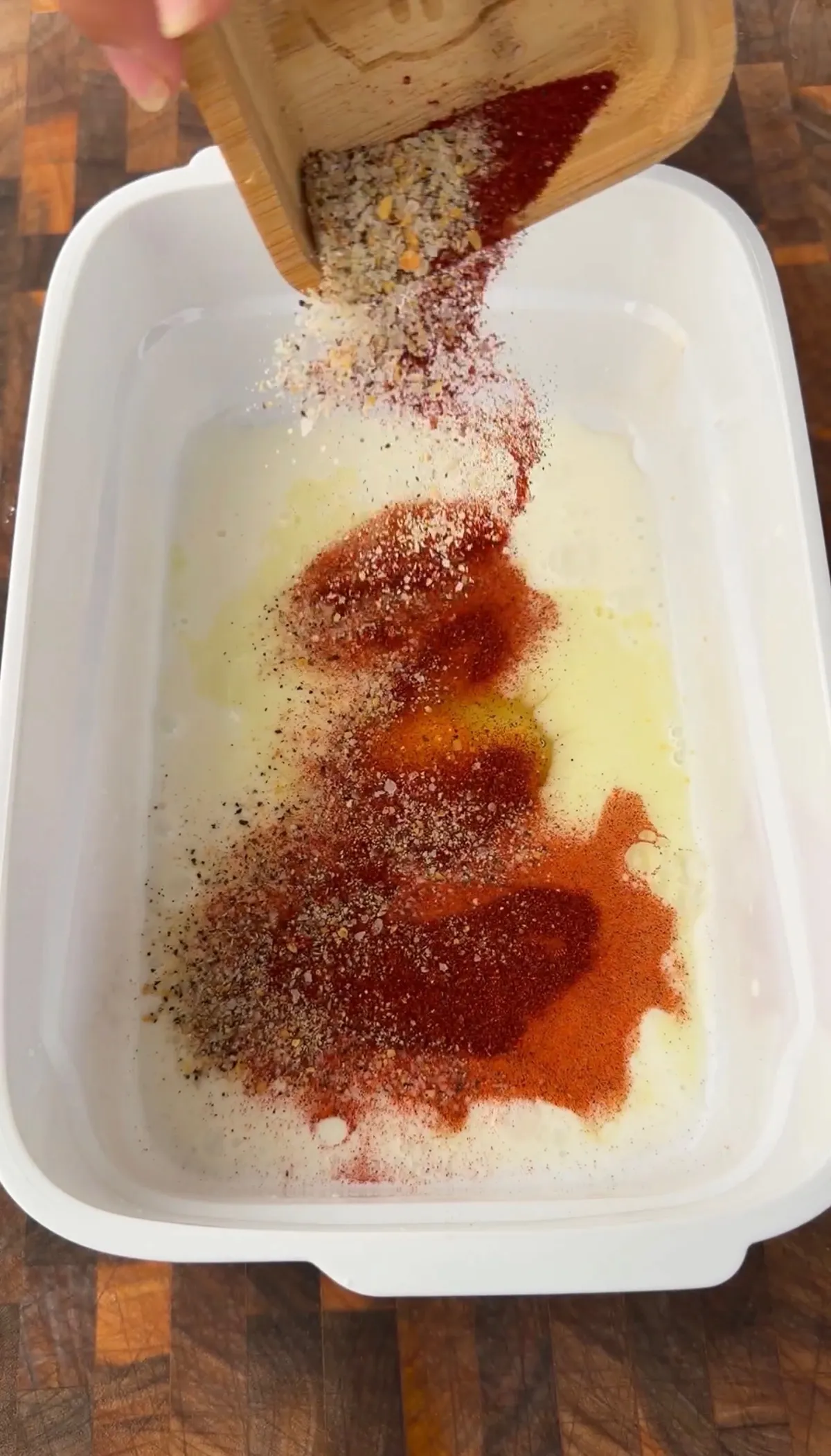 Pouring red spices and seasoning from wooden board into buttermilk marinade in white container for popcorn chicken