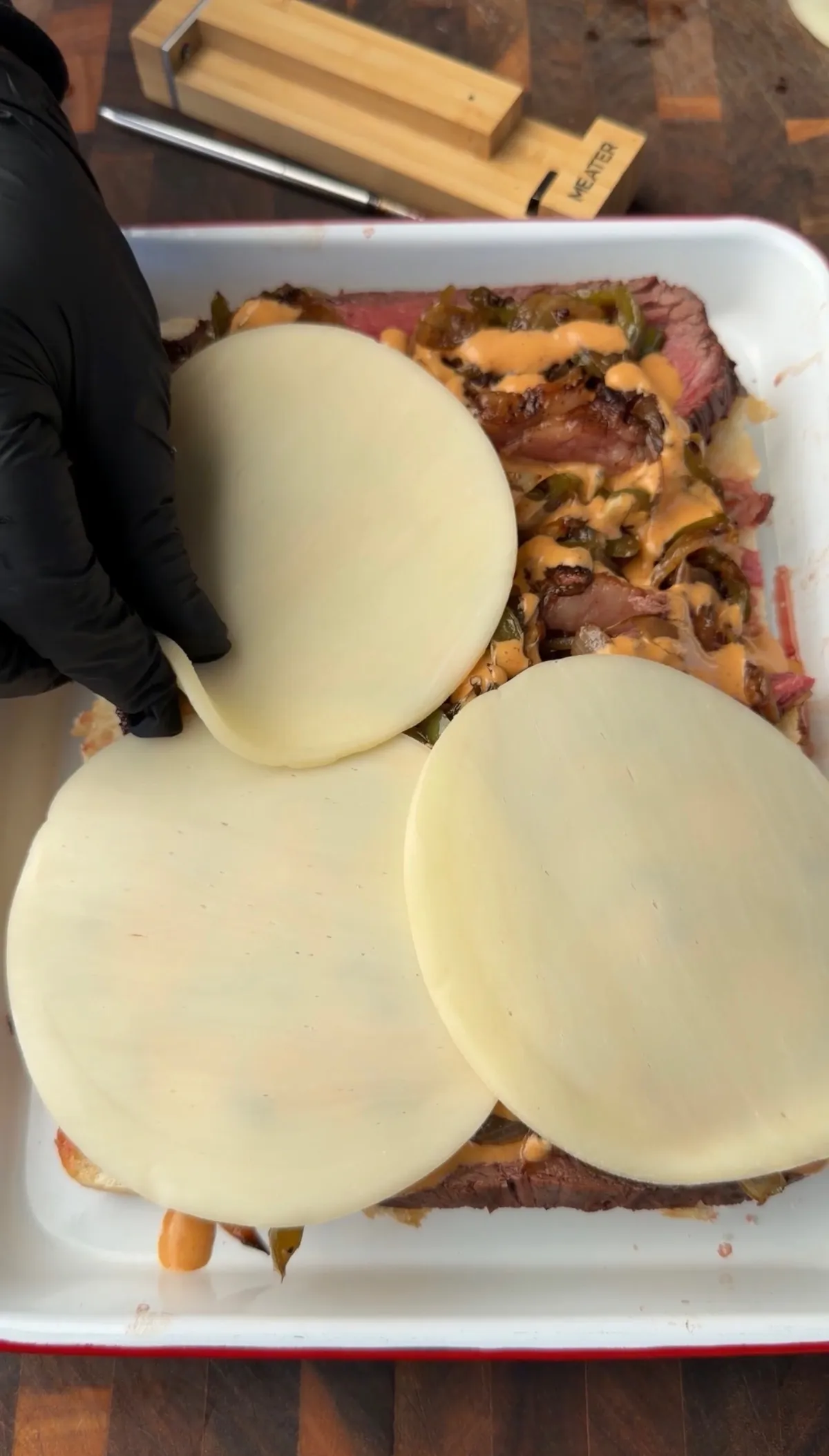 Provolone cheese slices being layered over picanha, sautéed peppers and onions, and chipotle mayo on Hawaiian roll sliders in a baking dish