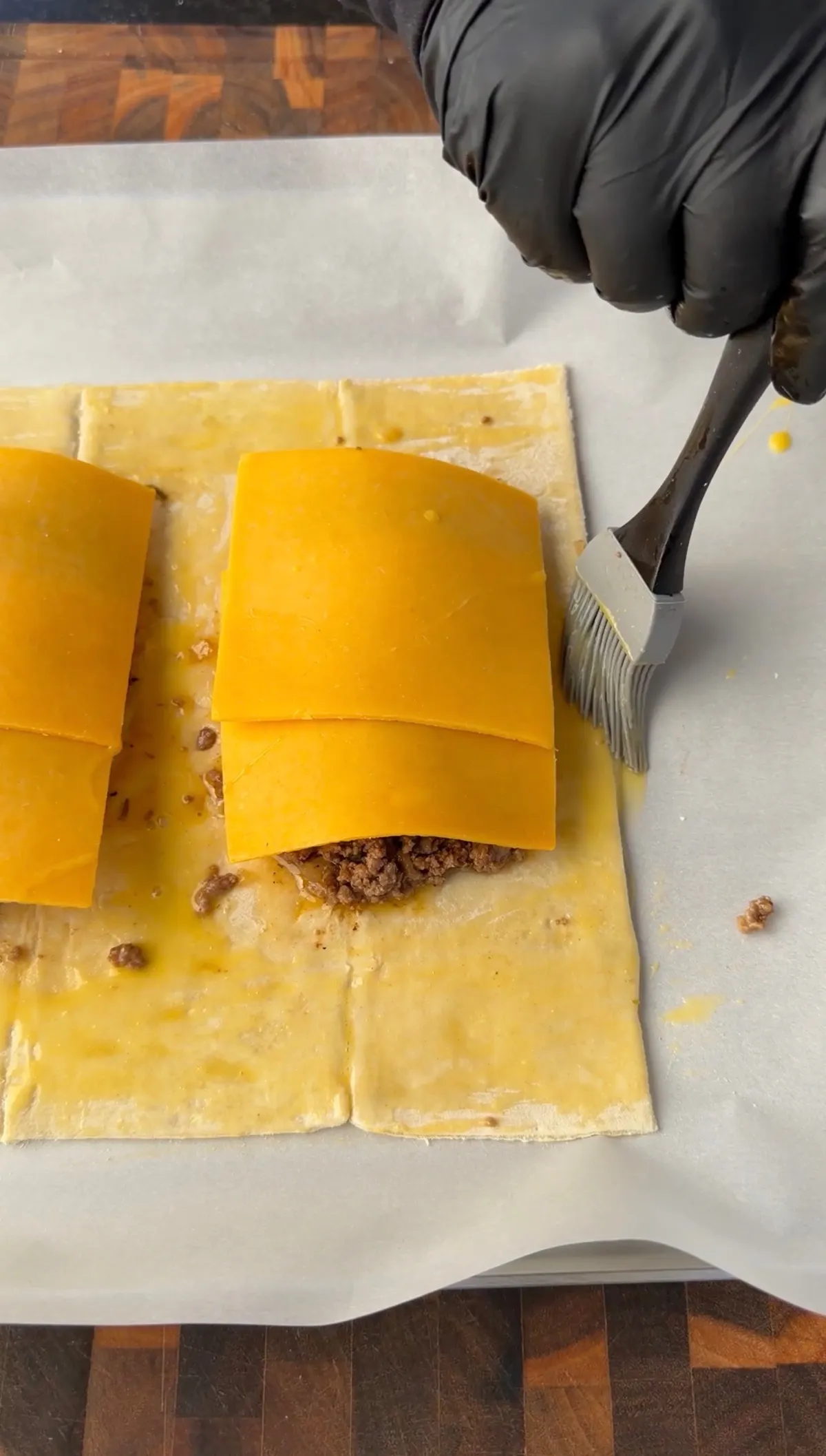 Assembling homemade cheeseburger hot pockets with cheddar cheese and ground beef on puff pastry, brushed with egg wash