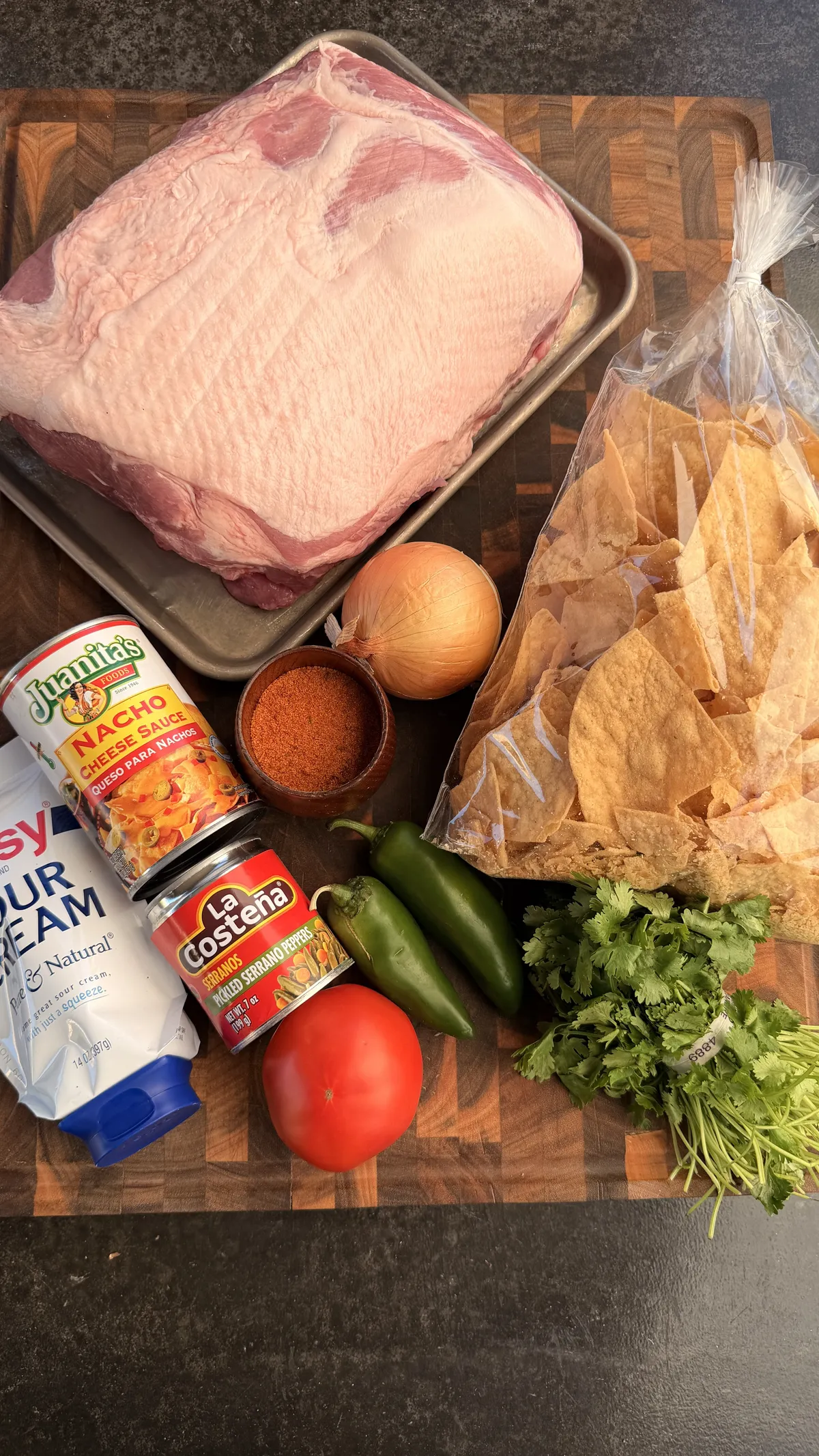 Flat lay of tailgating pulled pork nachos ingredients: bone-in pork shoulder, tortilla chips, nacho cheese sauce, sour cream, pickled serranos, jalapeños, tomato, onion, cilantro, and BBQ rub