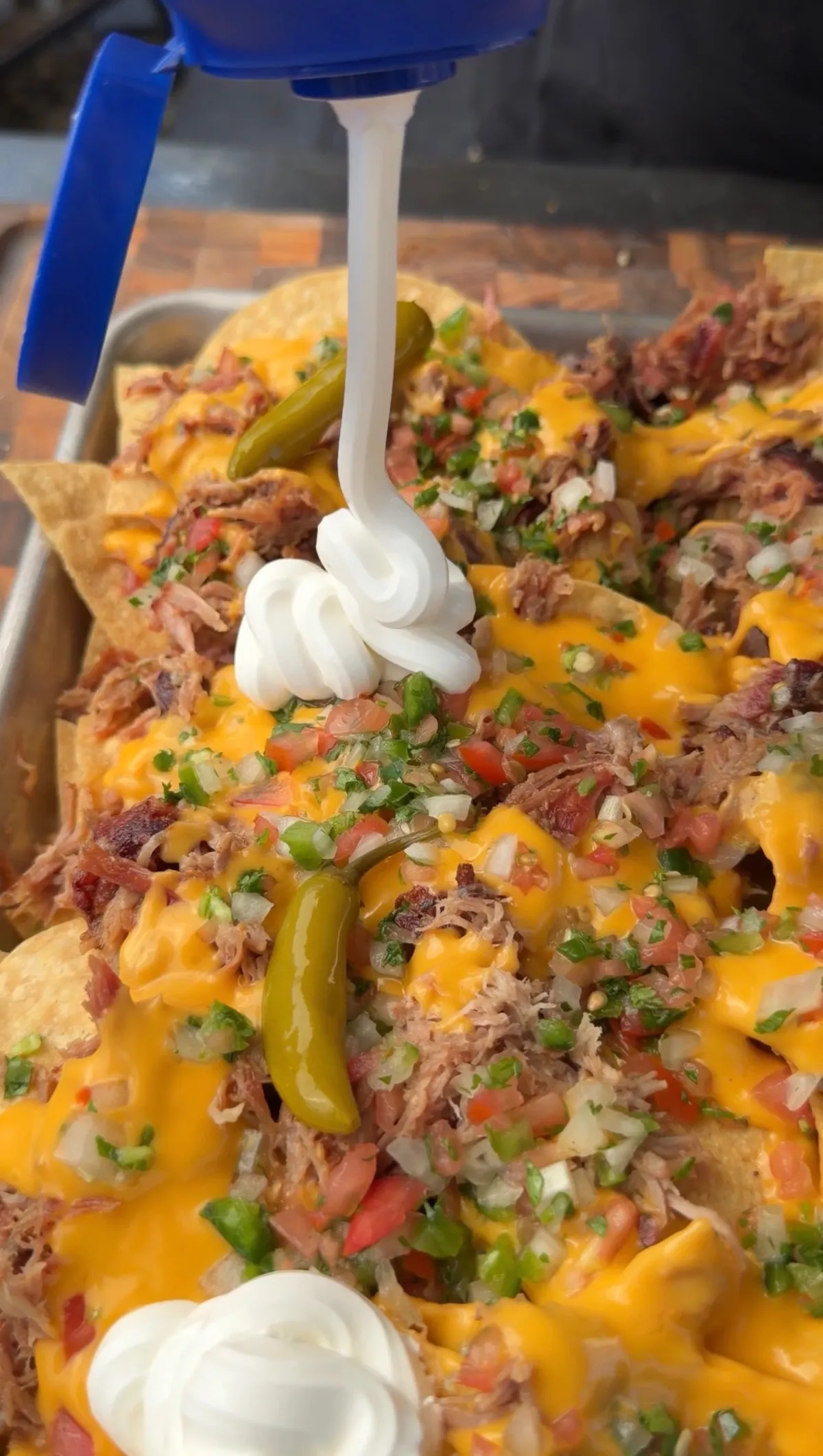 Tailgating pulled pork nachos topped with nacho cheese sauce, pico de gallo, pickled serranos, and sour cream being dolloped on top