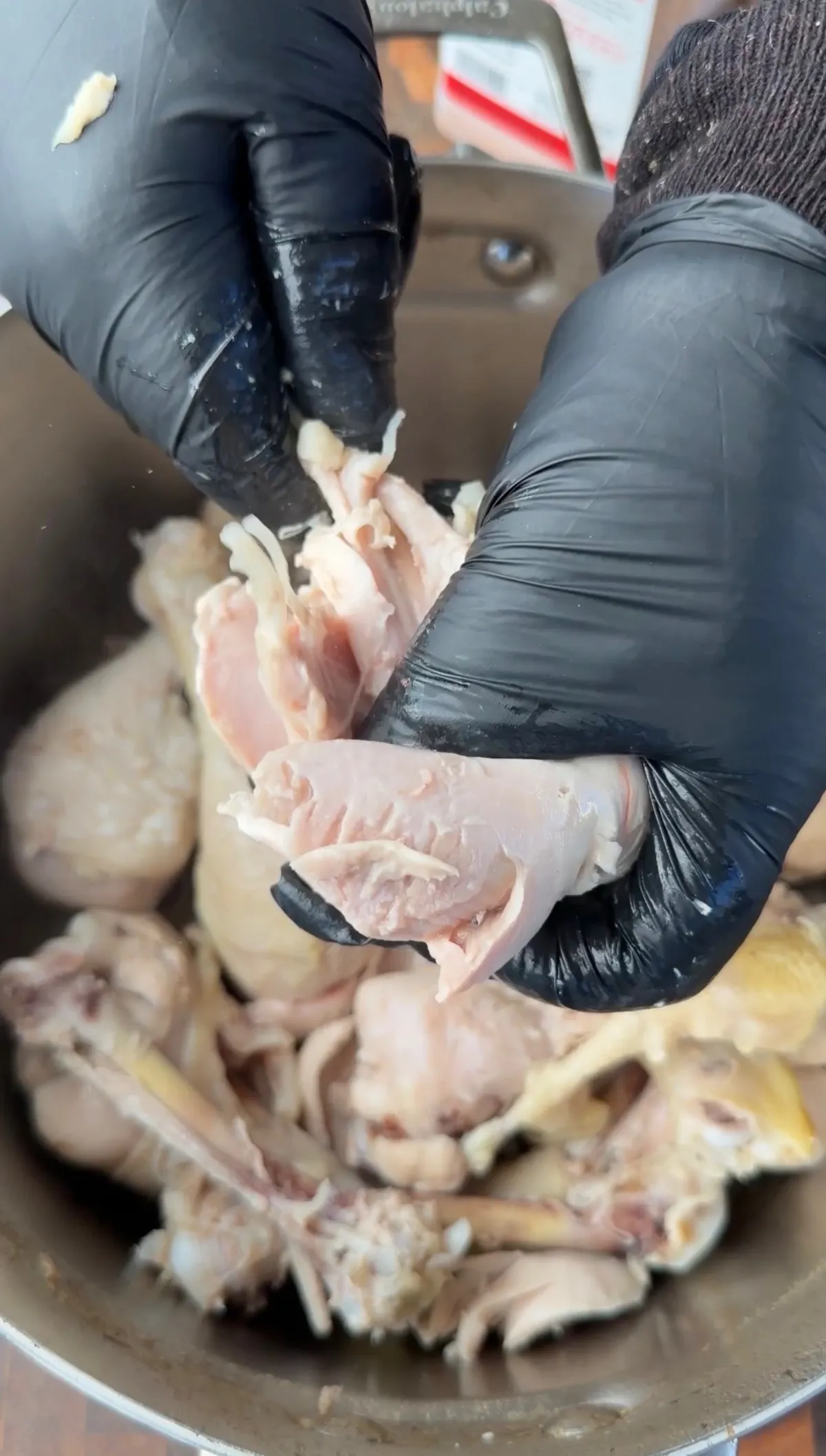 Gloved hands pulling shredded chicken meat from boiled drumstick bones over a large pot of cooked chicken pieces