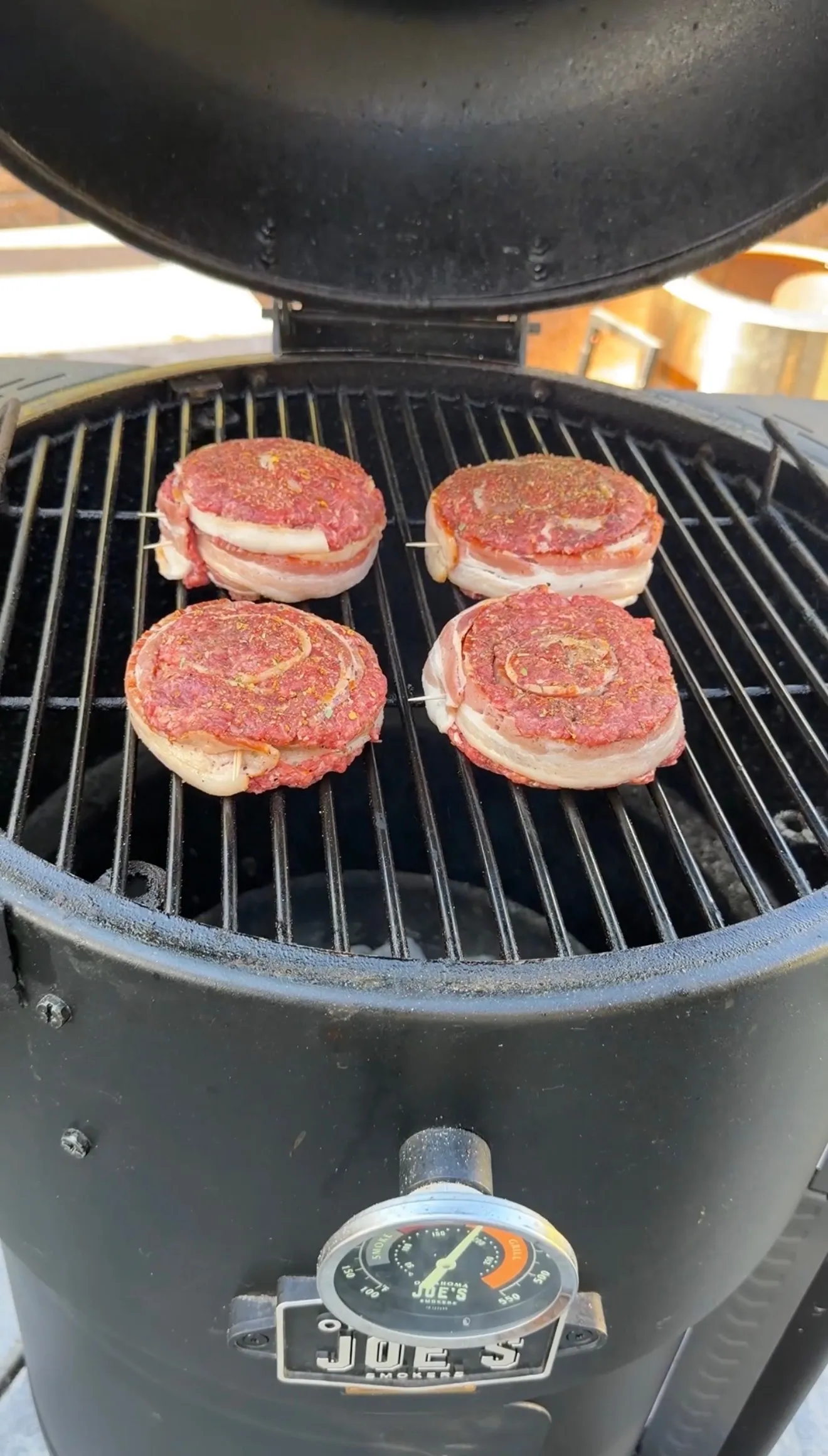 Four raw bacon-wrapped burger pinwheels seasoned with spices on a Joe's Smokers kamado grill