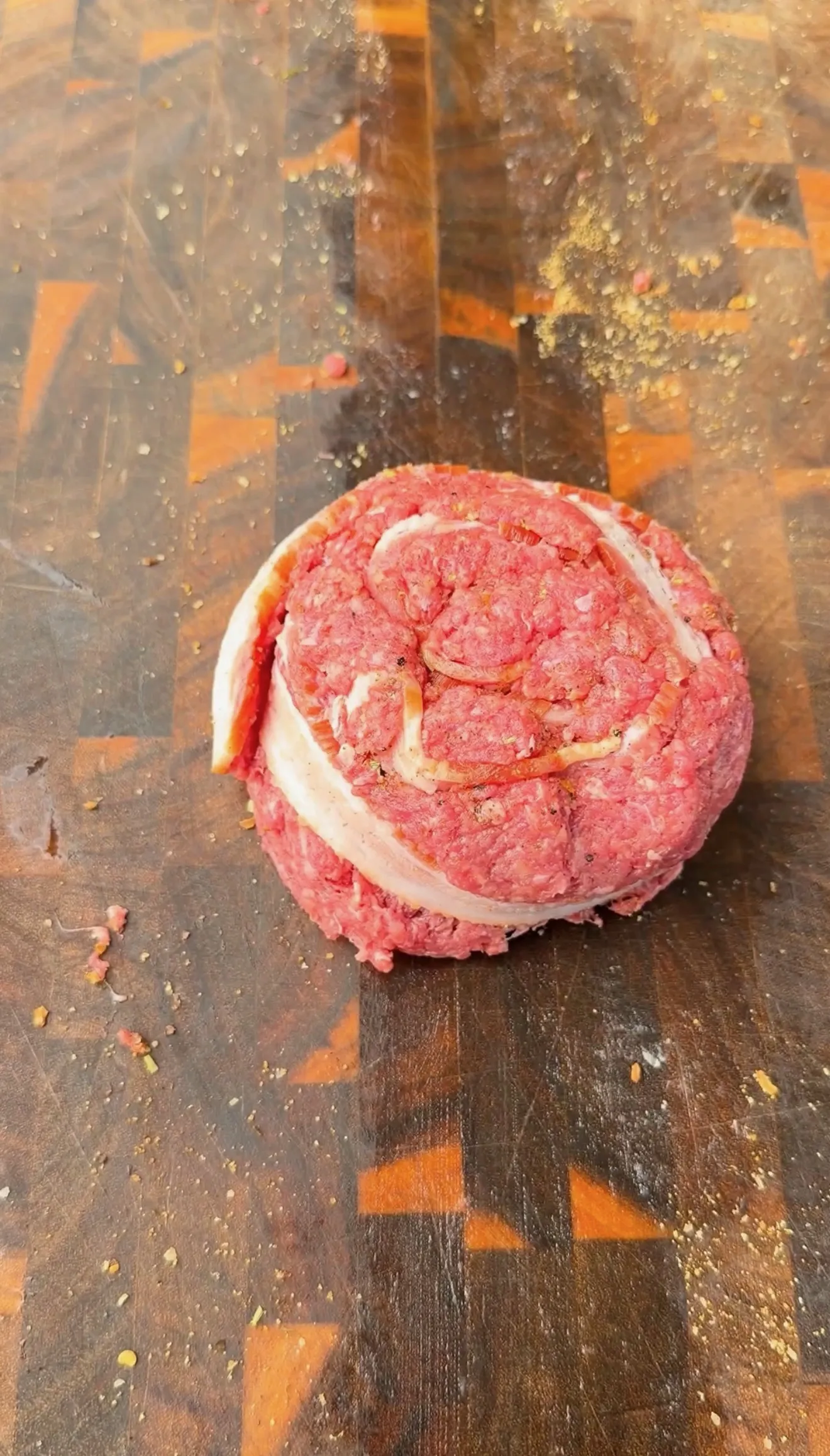 Raw ground beef burger patty wrapped with a strip of bacon on a seasoned wooden cutting board
