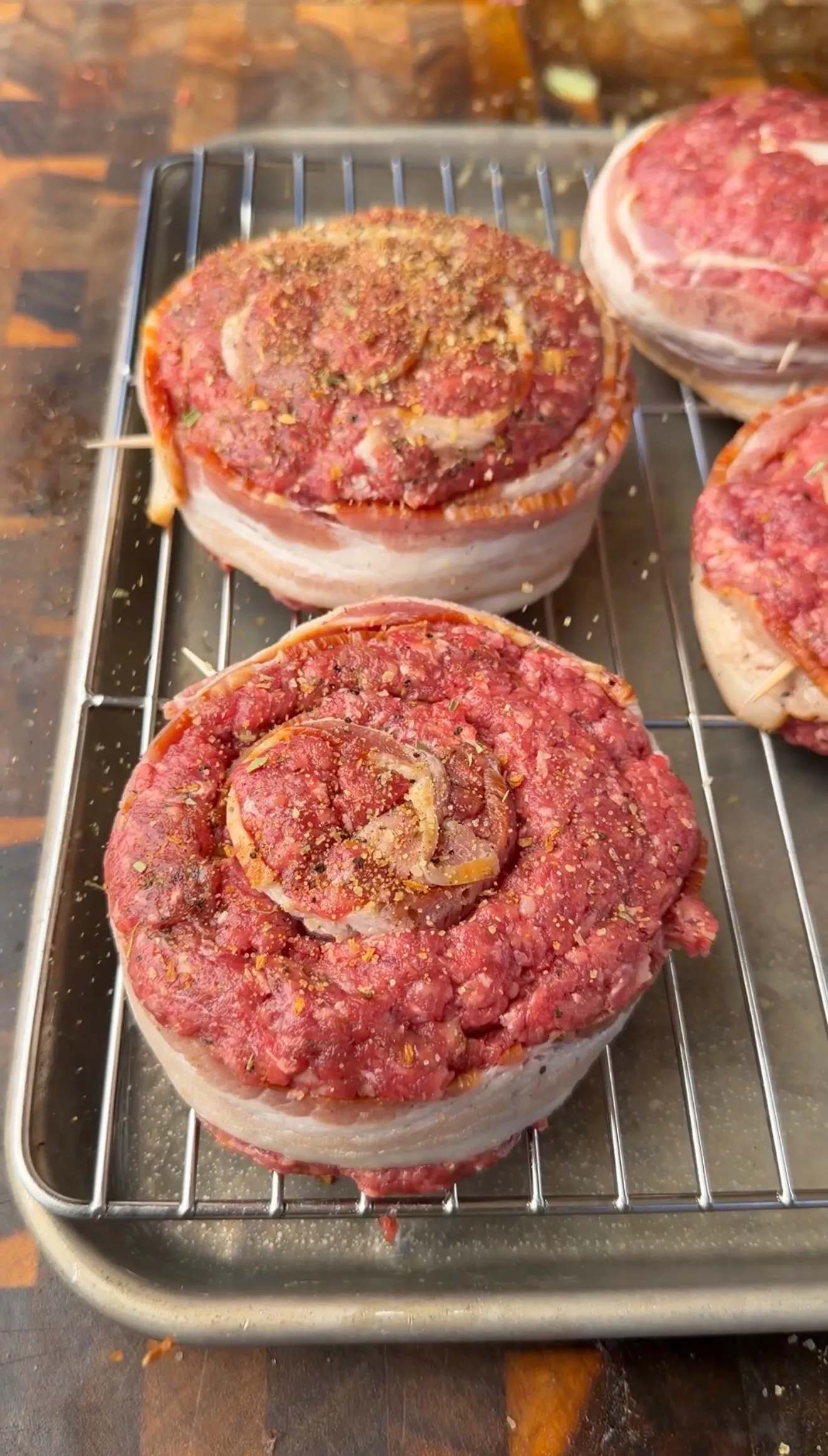 Raw bacon-wrapped ground beef pinwheel burgers seasoned with spices on a wire baking rack