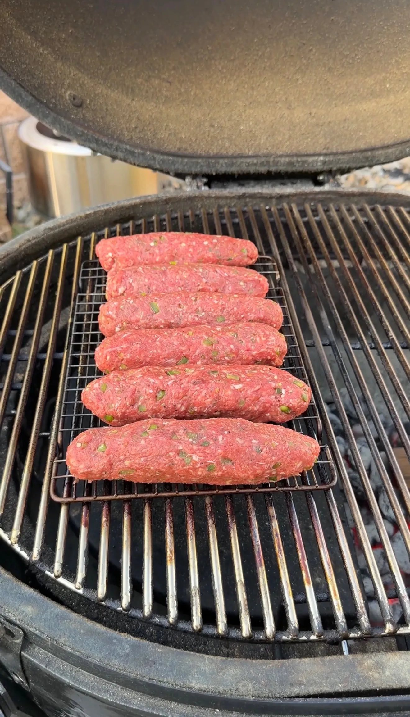 Six raw seasoned cheese stuffed burger dogs placed on a wire rack over charcoal grill grates