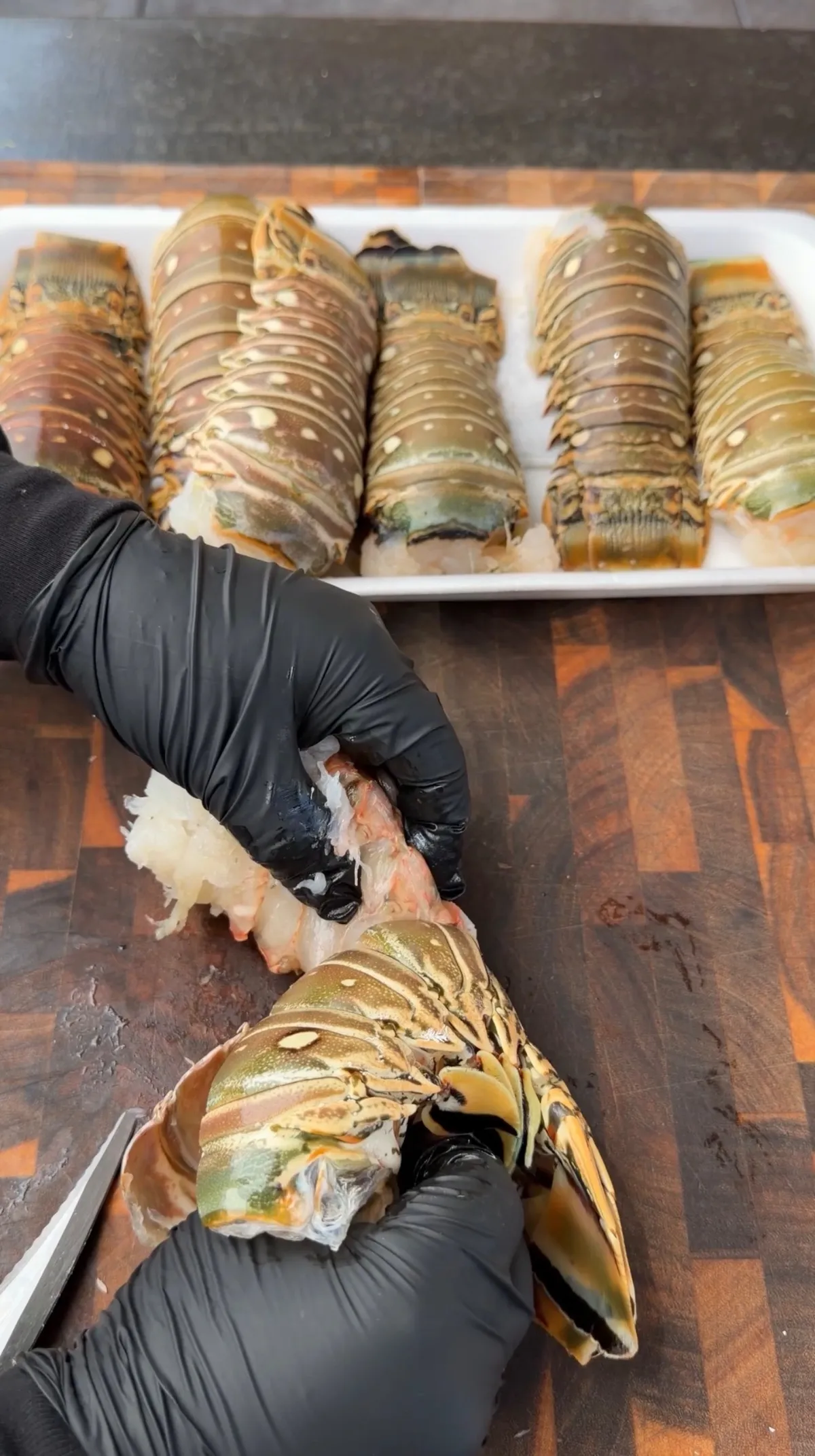 Hands in black gloves removing lobster meat from shell on wooden cutting board, with multiple raw lobster tails in background