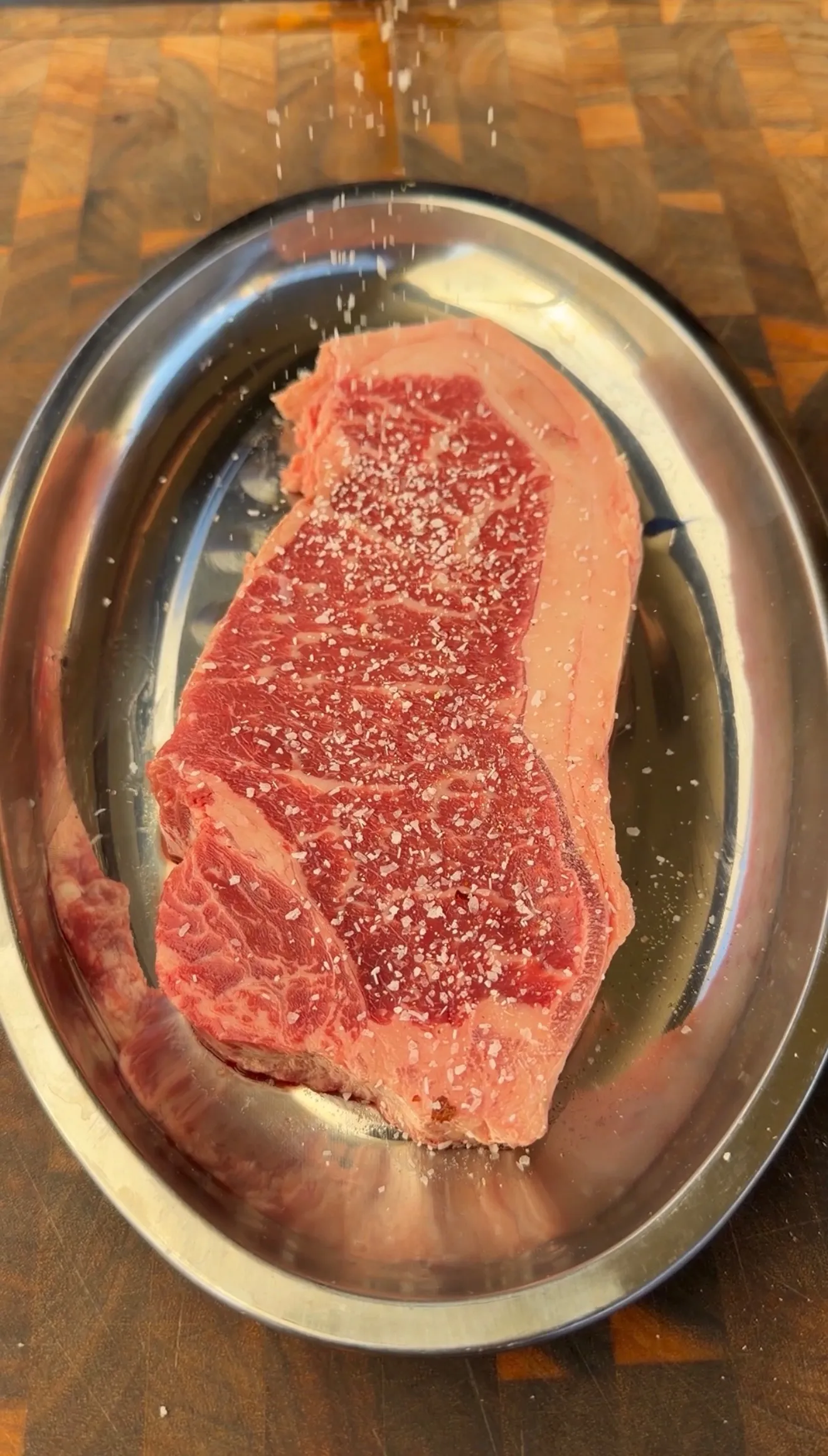 Raw New York strip steak being seasoned with coarse salt on a silver oval platter atop a wooden cutting board