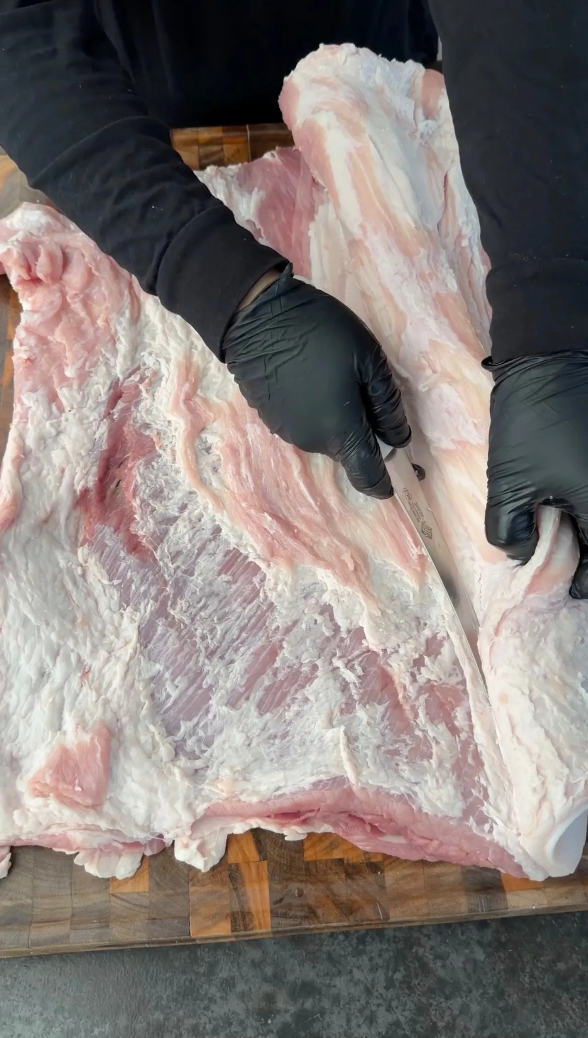 Gloved hands trimming a large raw pork belly slab with a knife on a wooden cutting board