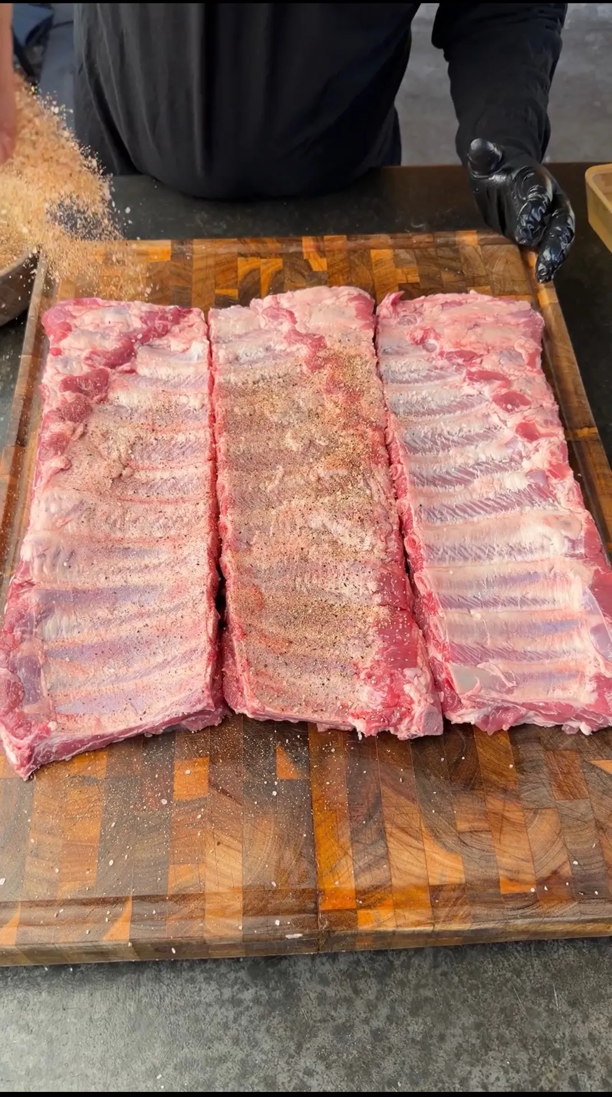 Three racks of raw pork ribs being seasoned with dry rub spice blend on a wooden butcher block cutting board