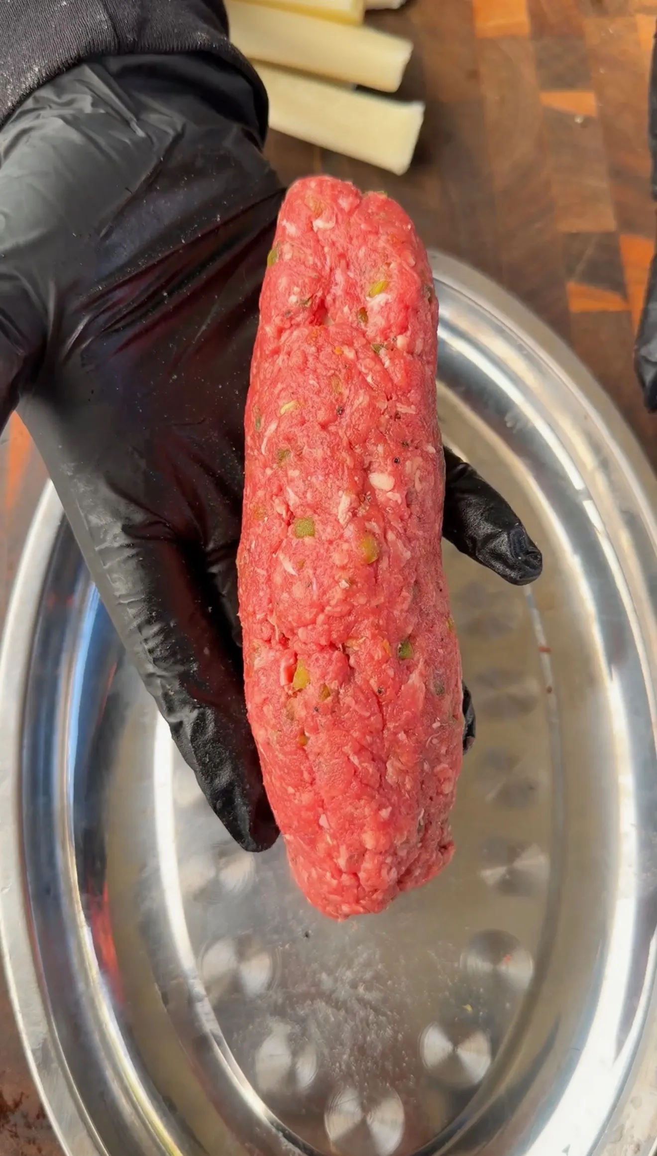 Gloved hand holding a seasoned raw ground beef log shaped for a cheese stuffed burger dog over a metal tray