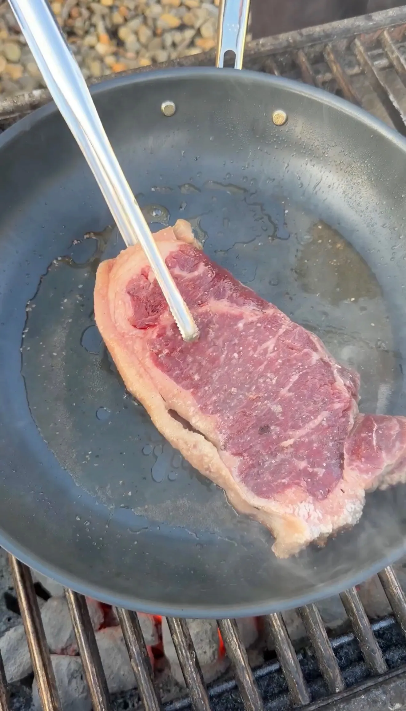 New York strip steak held with tongs in a dark skillet over a charcoal grill