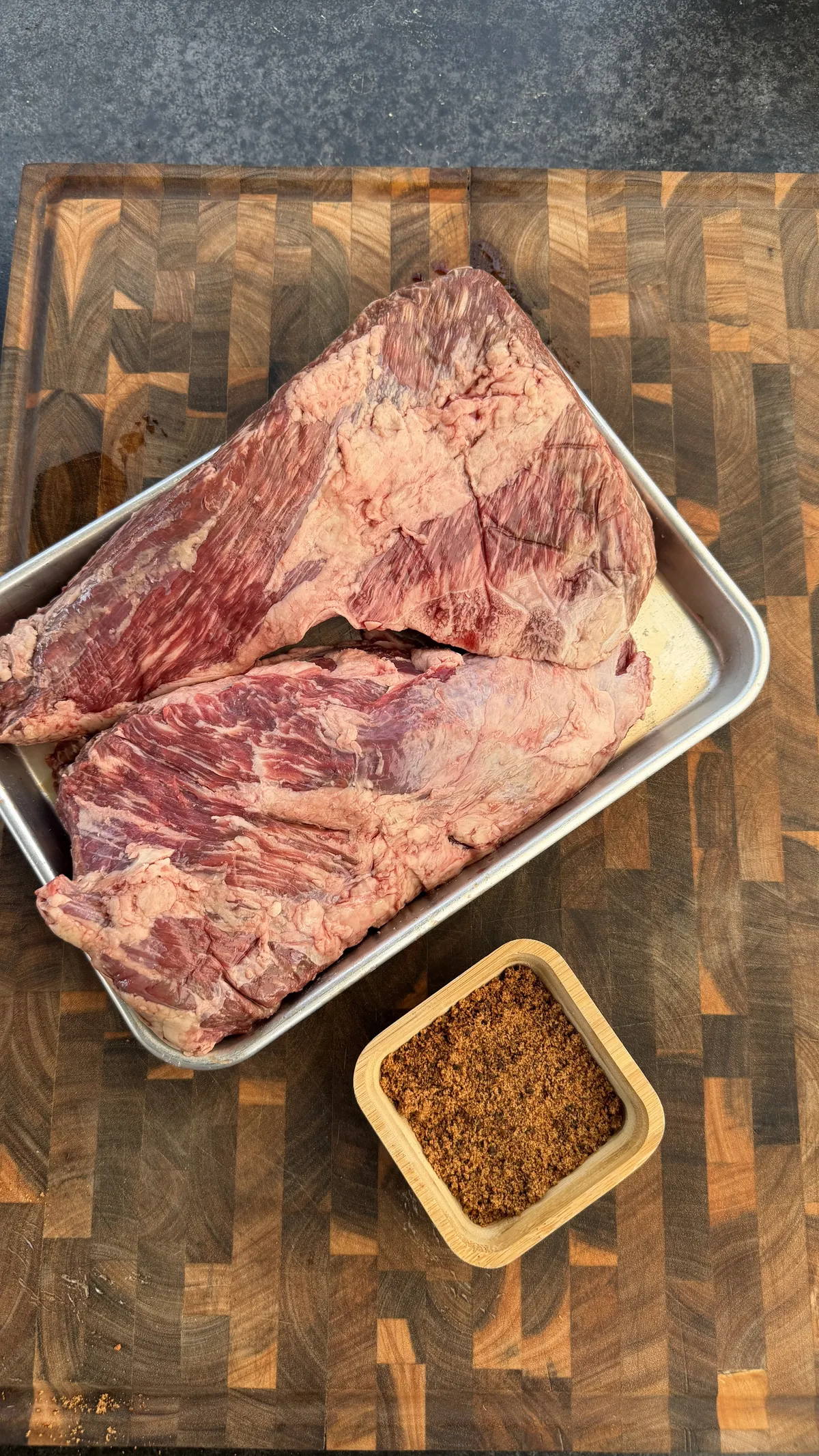 Raw tri-tip roast in a sheet pan with BBQ dry rub spice blend on an end-grain cutting board, prepped for trisket