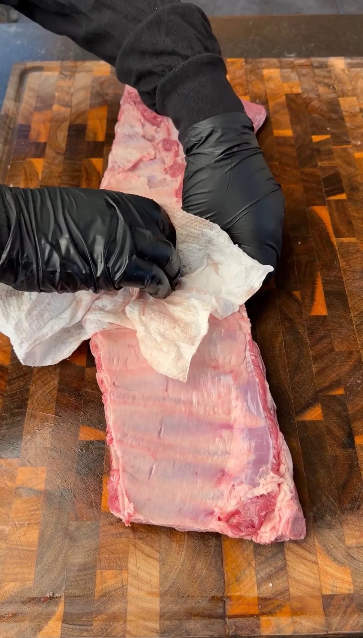 Gloved hands removing the silver skin membrane from a raw rack of pork ribs on a wooden cutting board