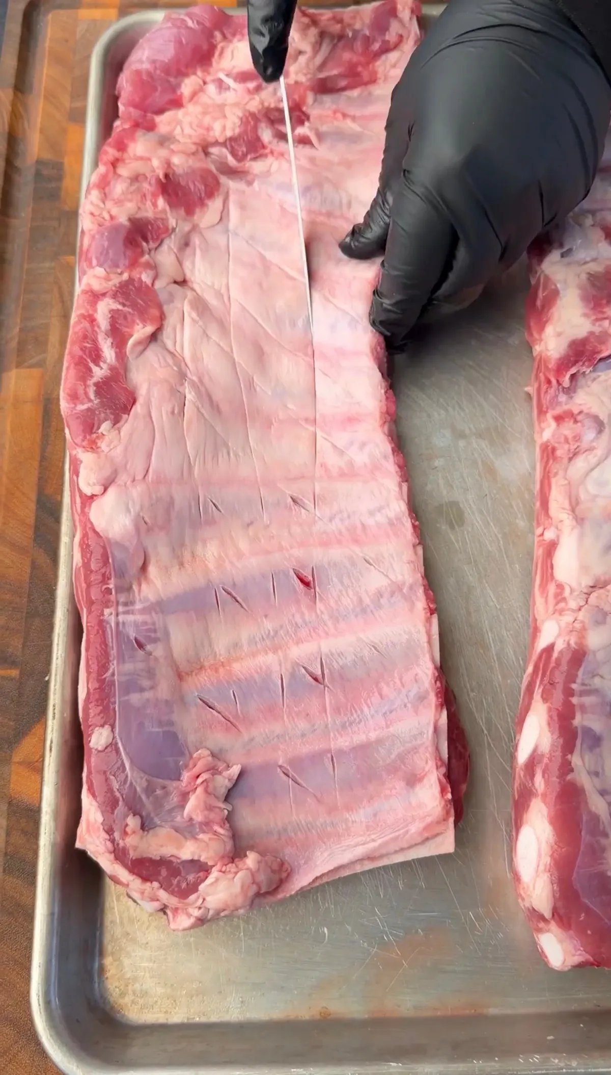 Hands in black gloves removing the silver skin membrane from a raw rack of St. Louis spare ribs on a baking sheet
