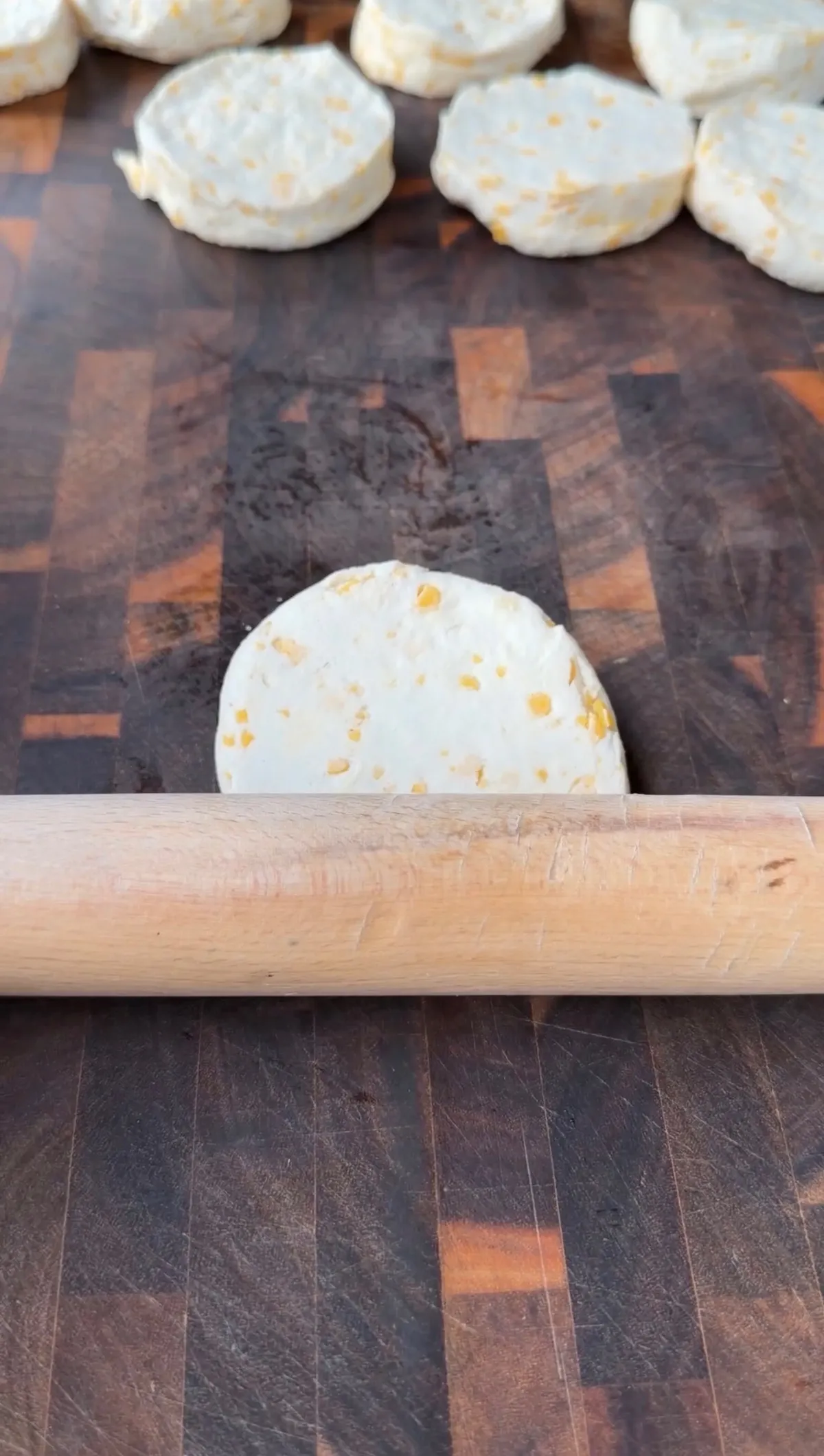 A wooden rolling pin flattening a round cheese biscuit dough round on a butcher block cutting board