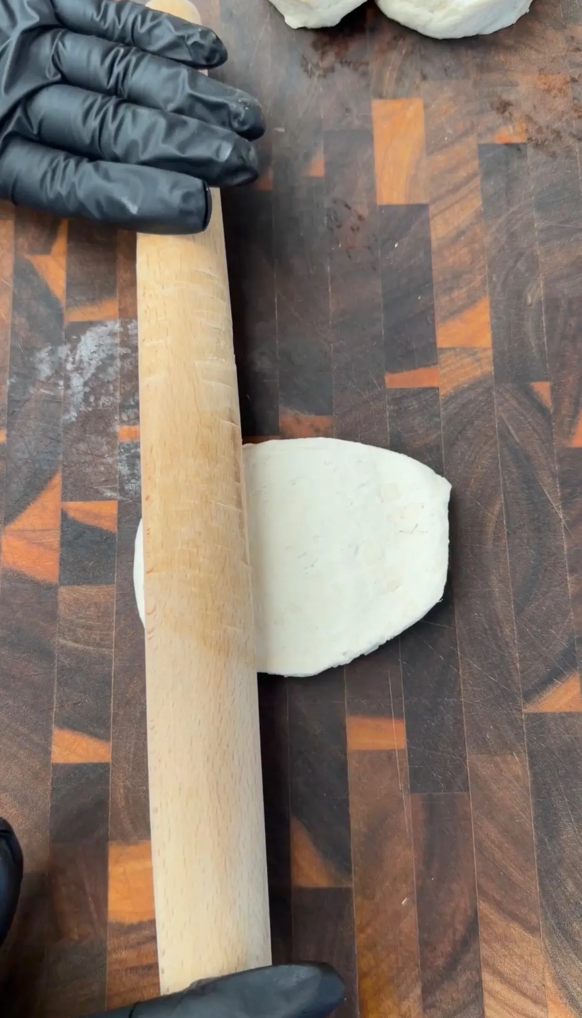 Hands in black gloves using a wooden rolling pin to flatten white dough on a wood cutting board for pizza bombs