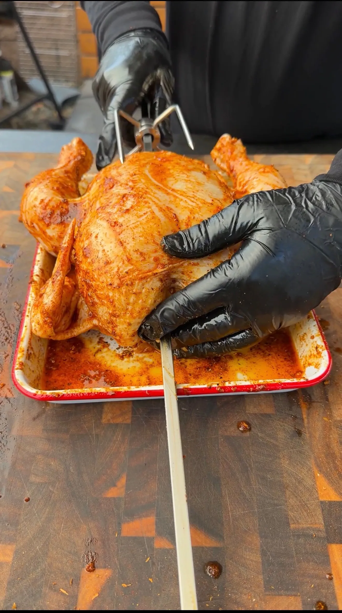 Whole Cajun-spiced chicken being mounted on a rotisserie spit rod in a roasting pan by gloved hands