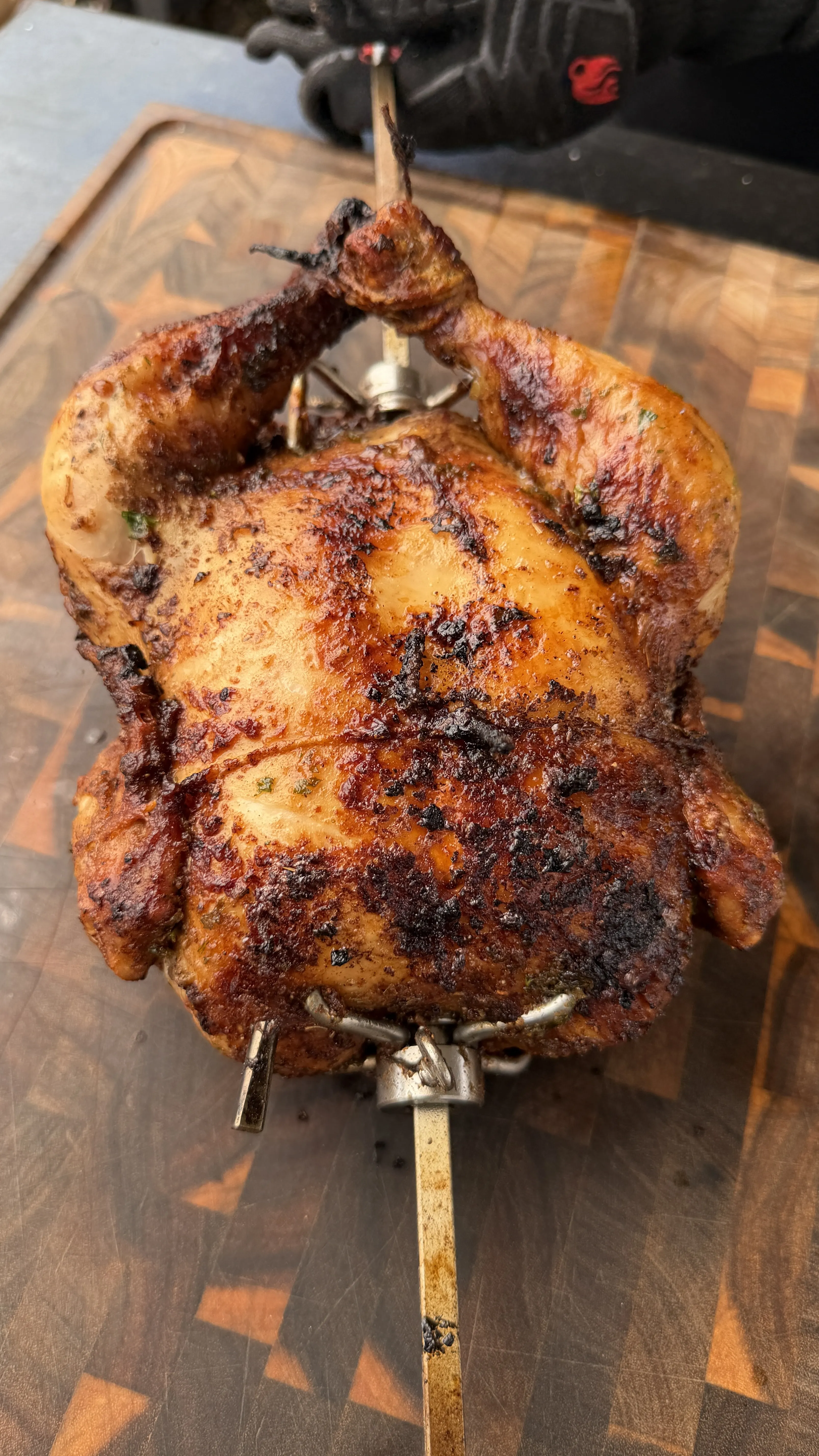 Whole Cajun butter rotisserie chicken on a spit rod, golden-brown and charred, resting on a wooden cutting board