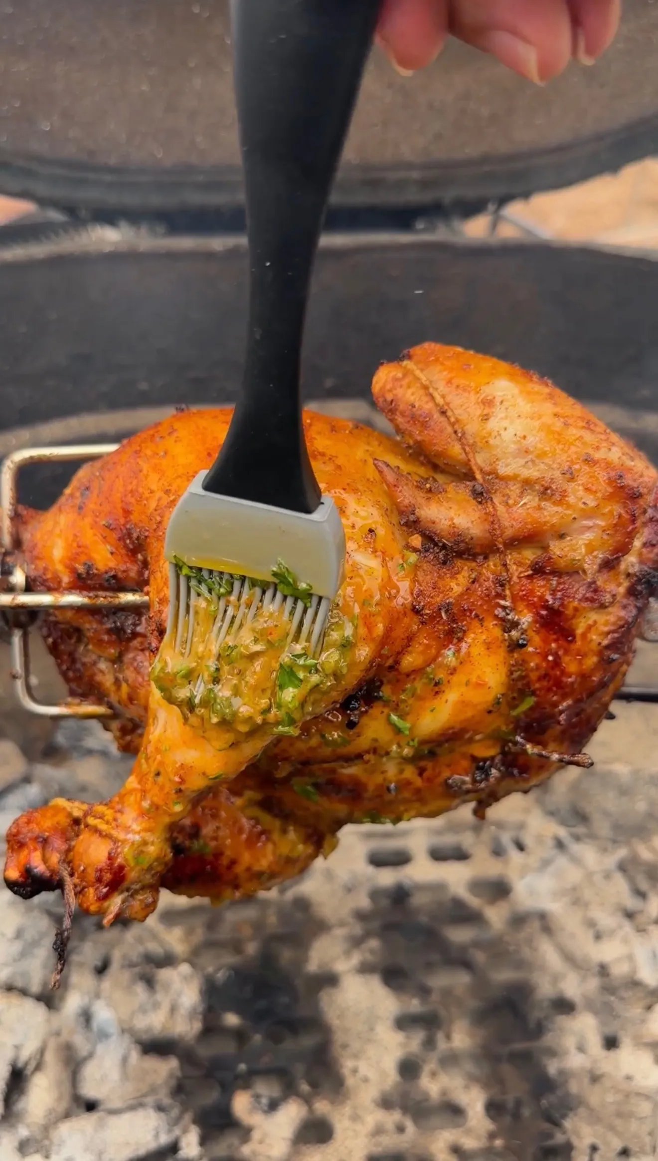 Whole rotisserie Cajun chicken being basted with herb butter sauce using a silicone brush on a charcoal grill