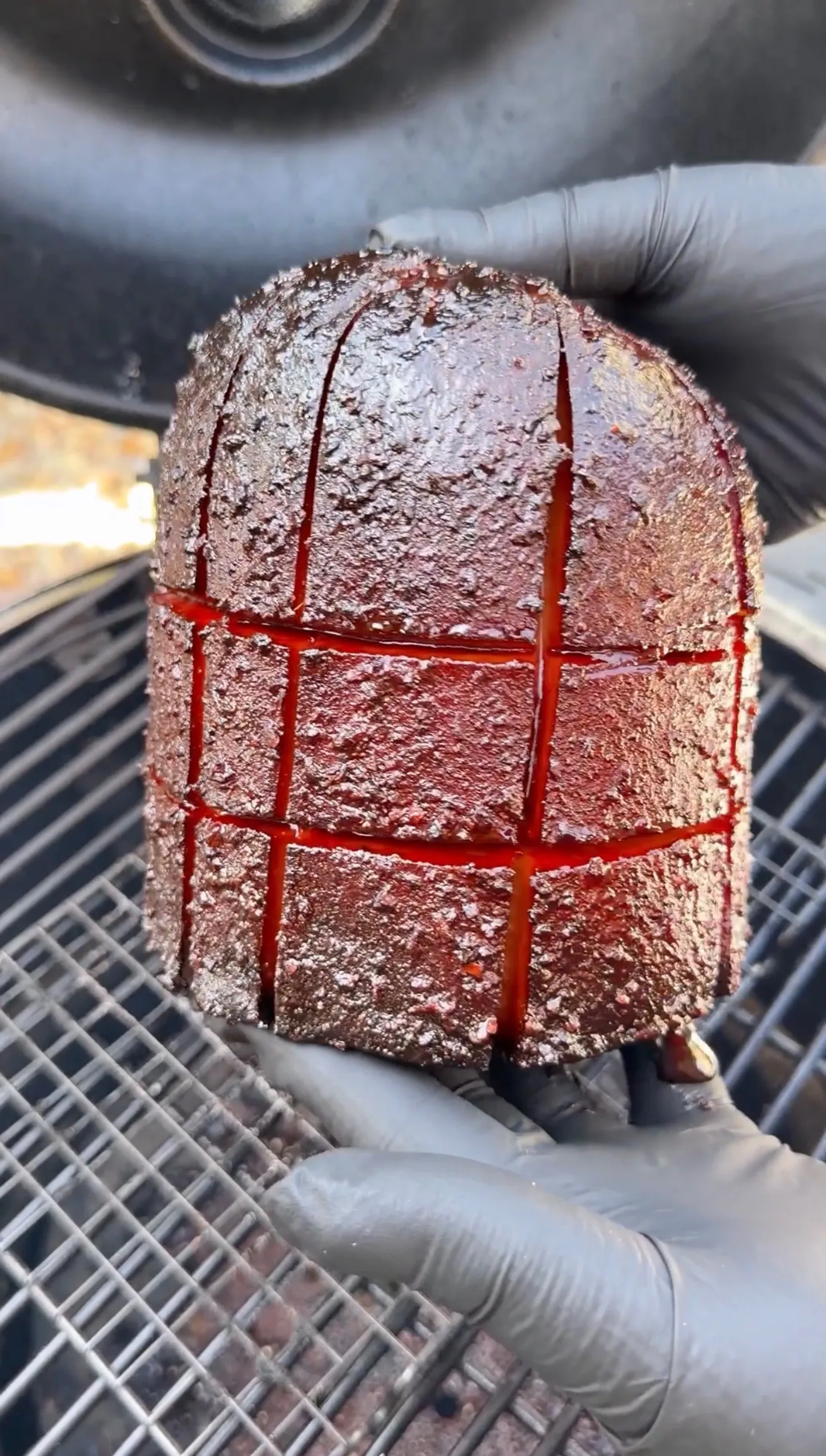 Hands holding a scored bologna chub with a dark bark crust and red glaze on a smoker grill grate