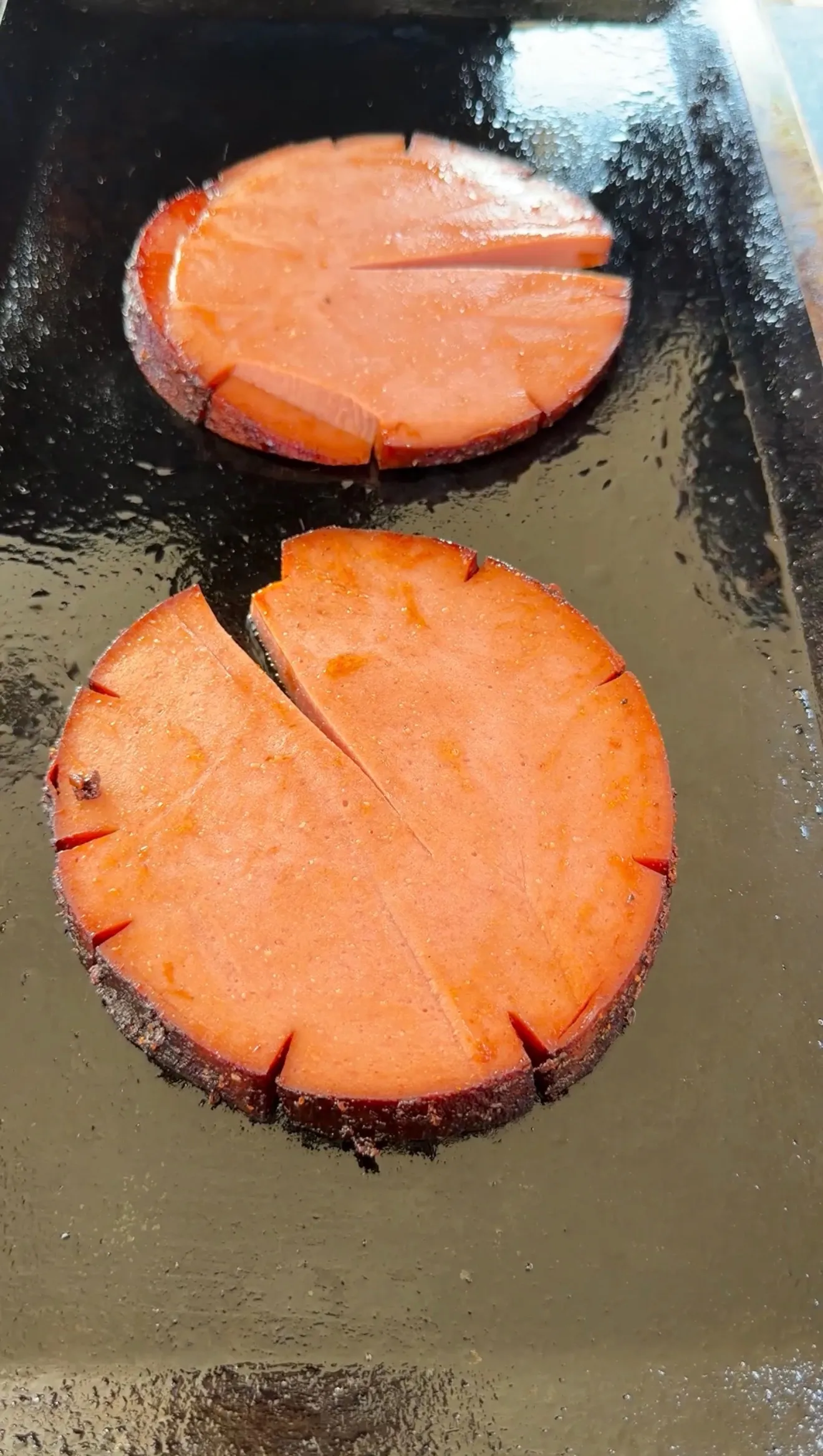 Two thick scored bologna slices with crosshatch cuts cooking on a greased cast iron griddle