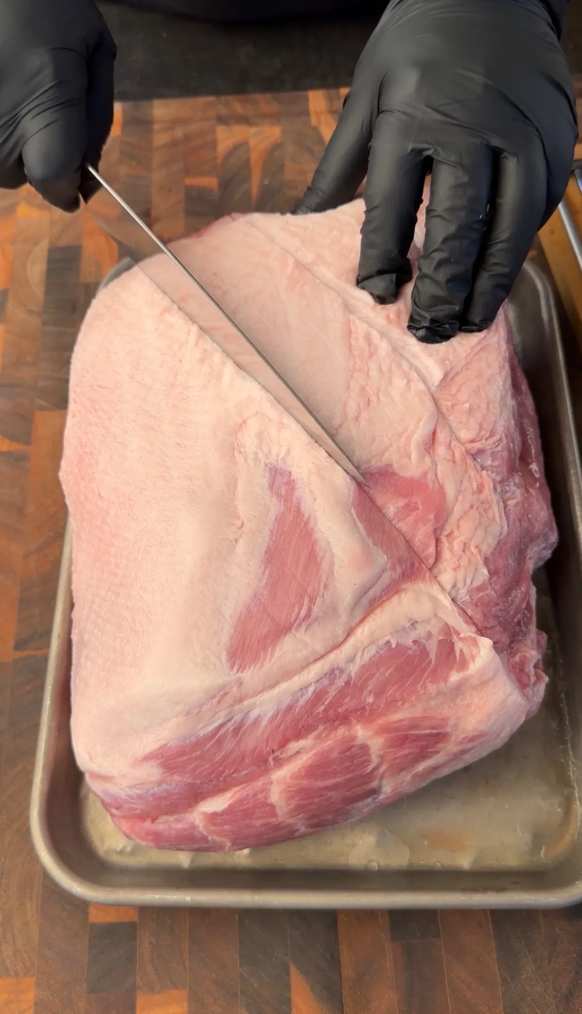 Gloved hands scoring the fat cap of a bone-in pork butt in a crosshatch pattern on a sheet tray