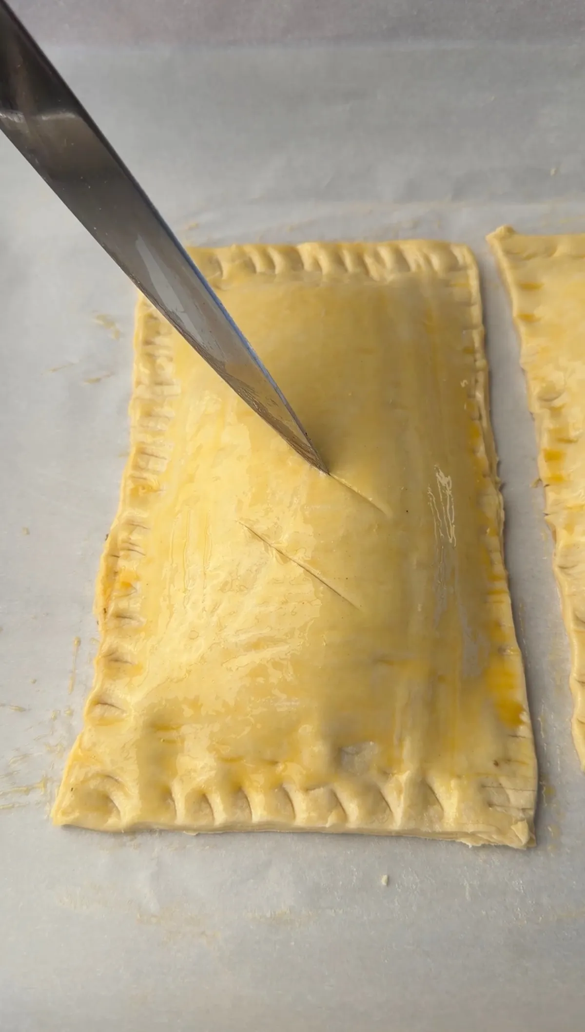 A knife scoring the top of an unbaked puff pastry cheeseburger hot pocket with crimped edges on parchment paper