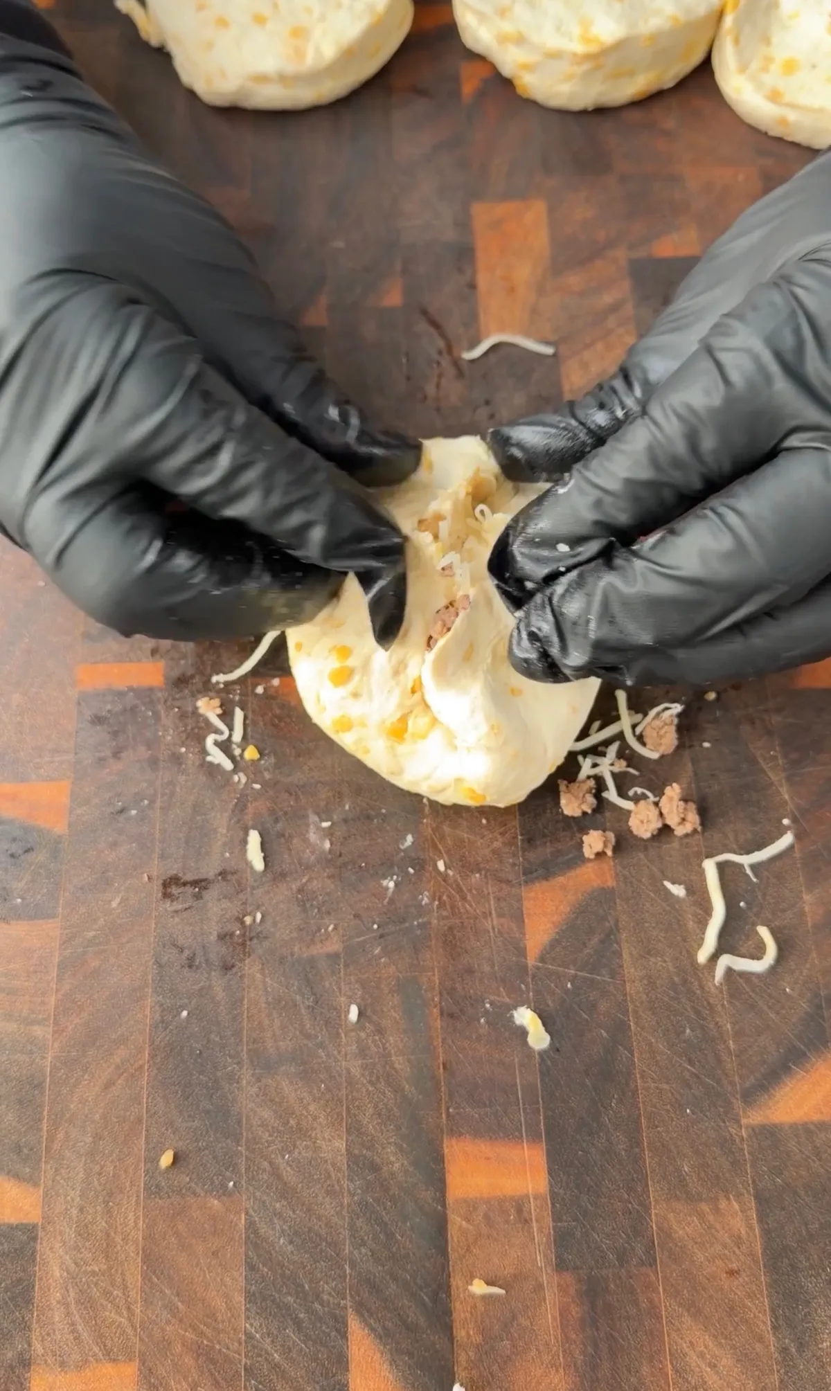 Hands in black gloves sealing cheeseburger filling inside biscuit dough on a wooden cutting board