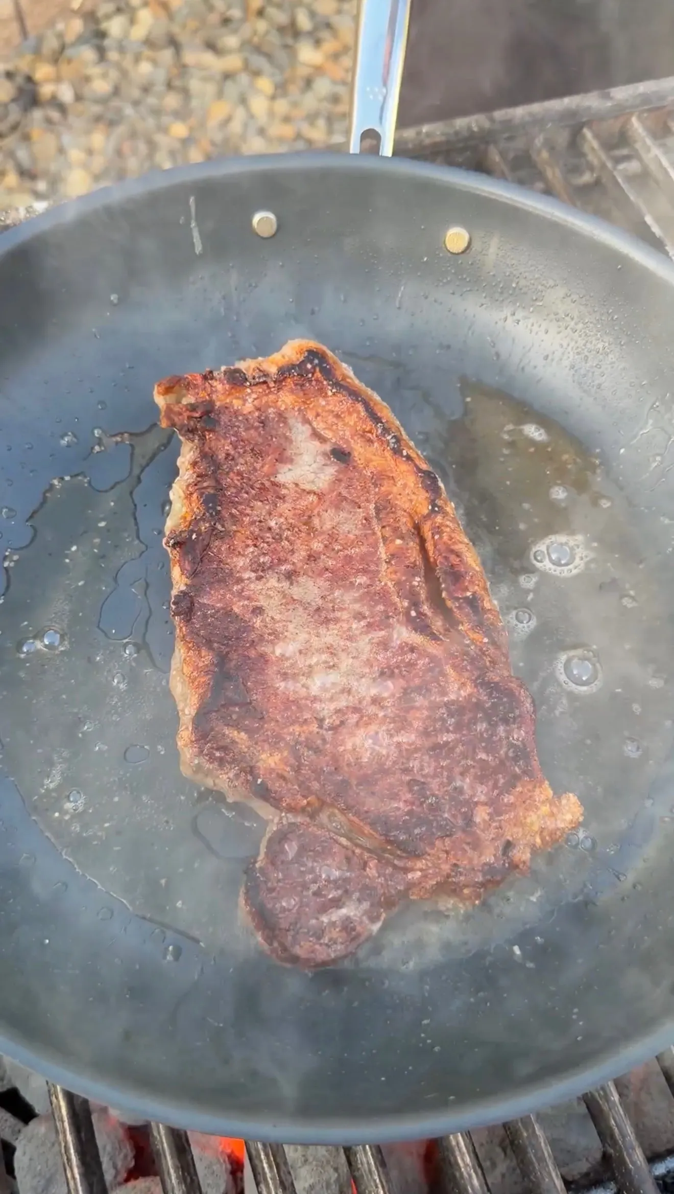 A seasoned steak with a golden-brown crust sizzling in a dark skillet over an open flame