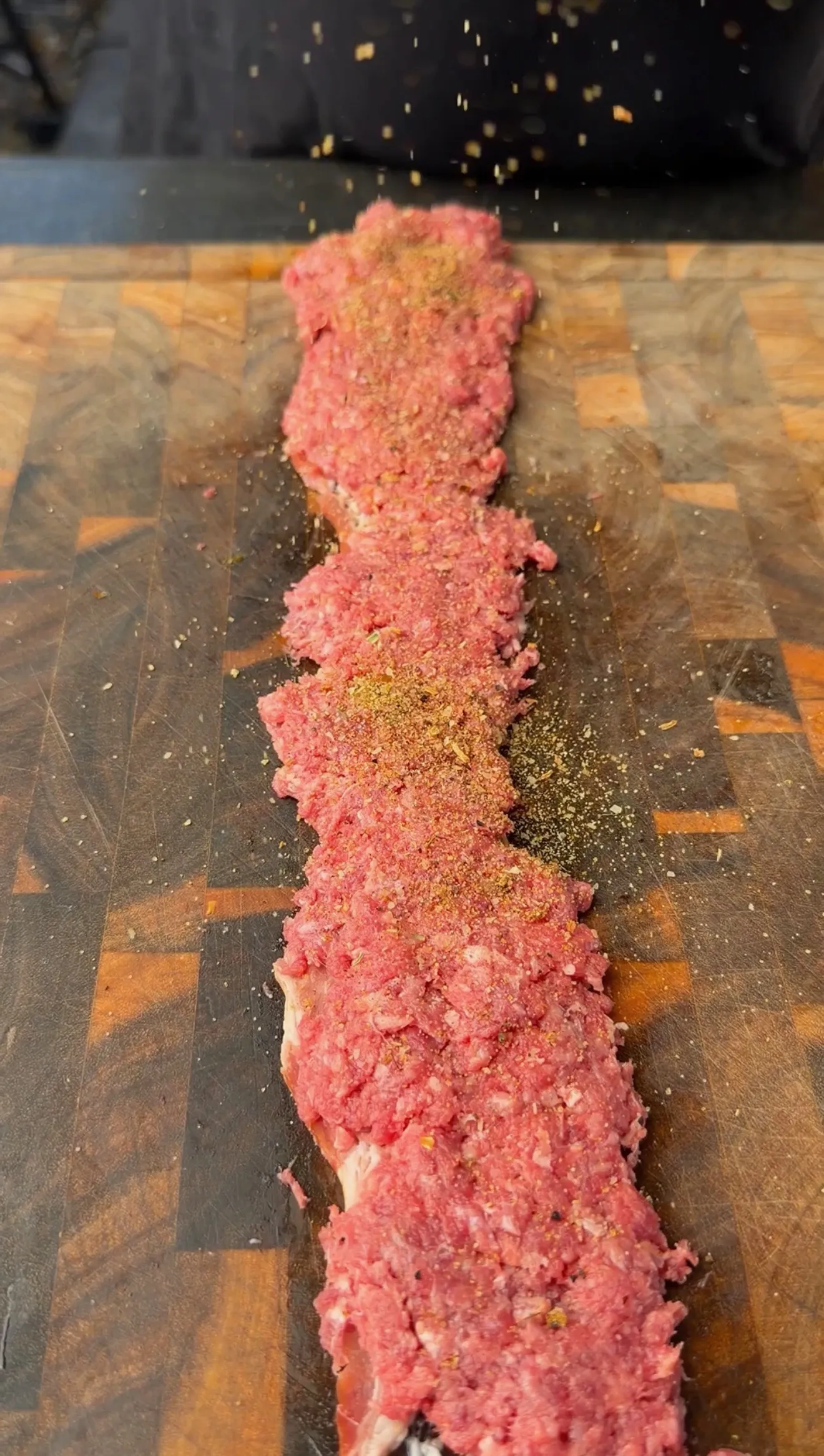 Raw seasoned ground beef spread in a long log shape on a wooden cutting board, sprinkled with spices