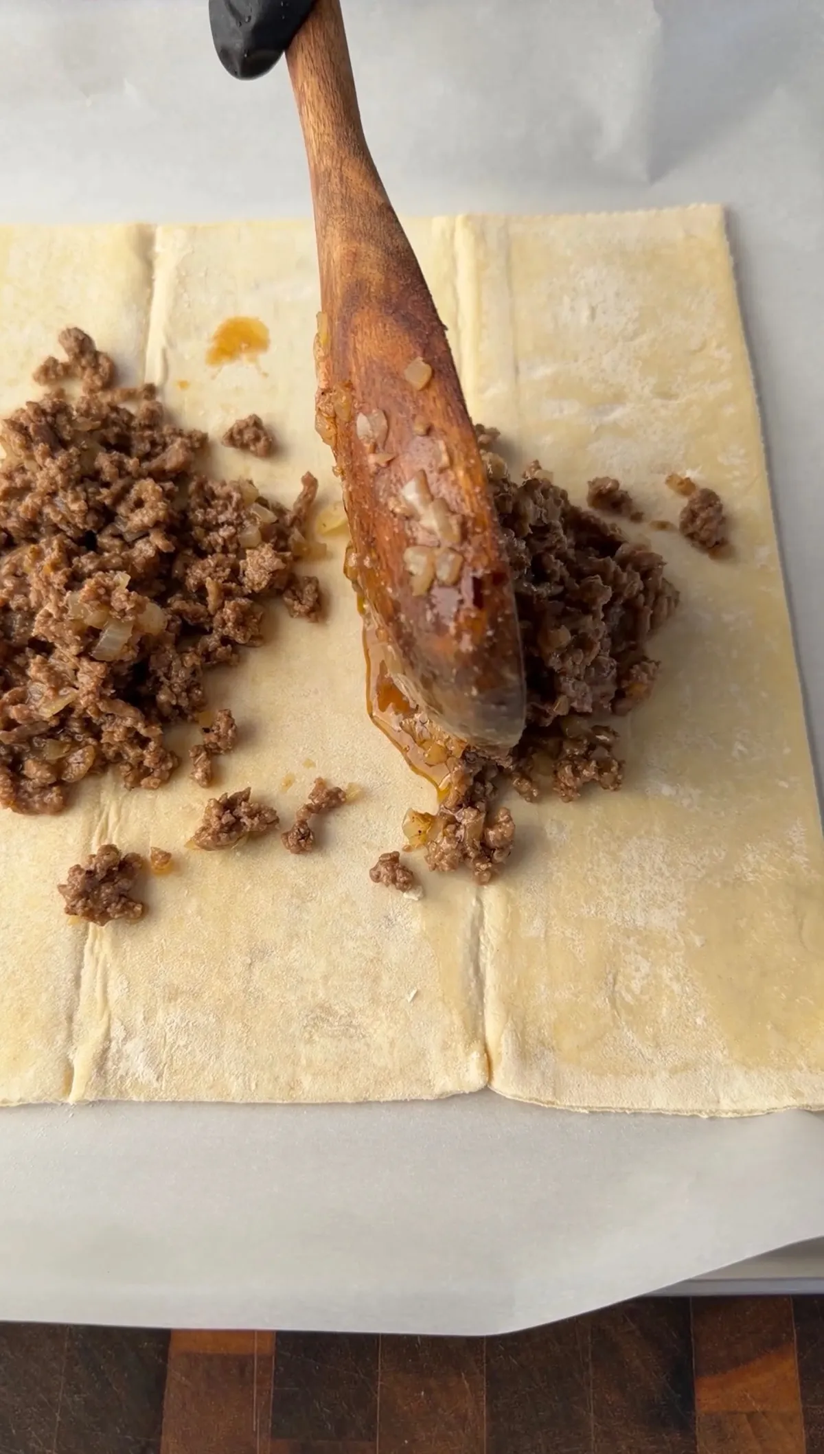 Seasoned ground beef mixture being spread onto flat puff pastry dough with a wooden spoon for cheeseburger hot pockets