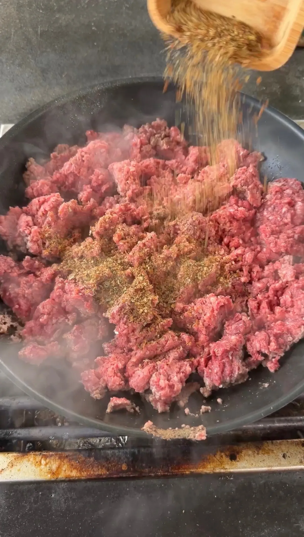 Spices being poured from a wooden bowl onto raw ground beef sizzling in a cast iron skillet