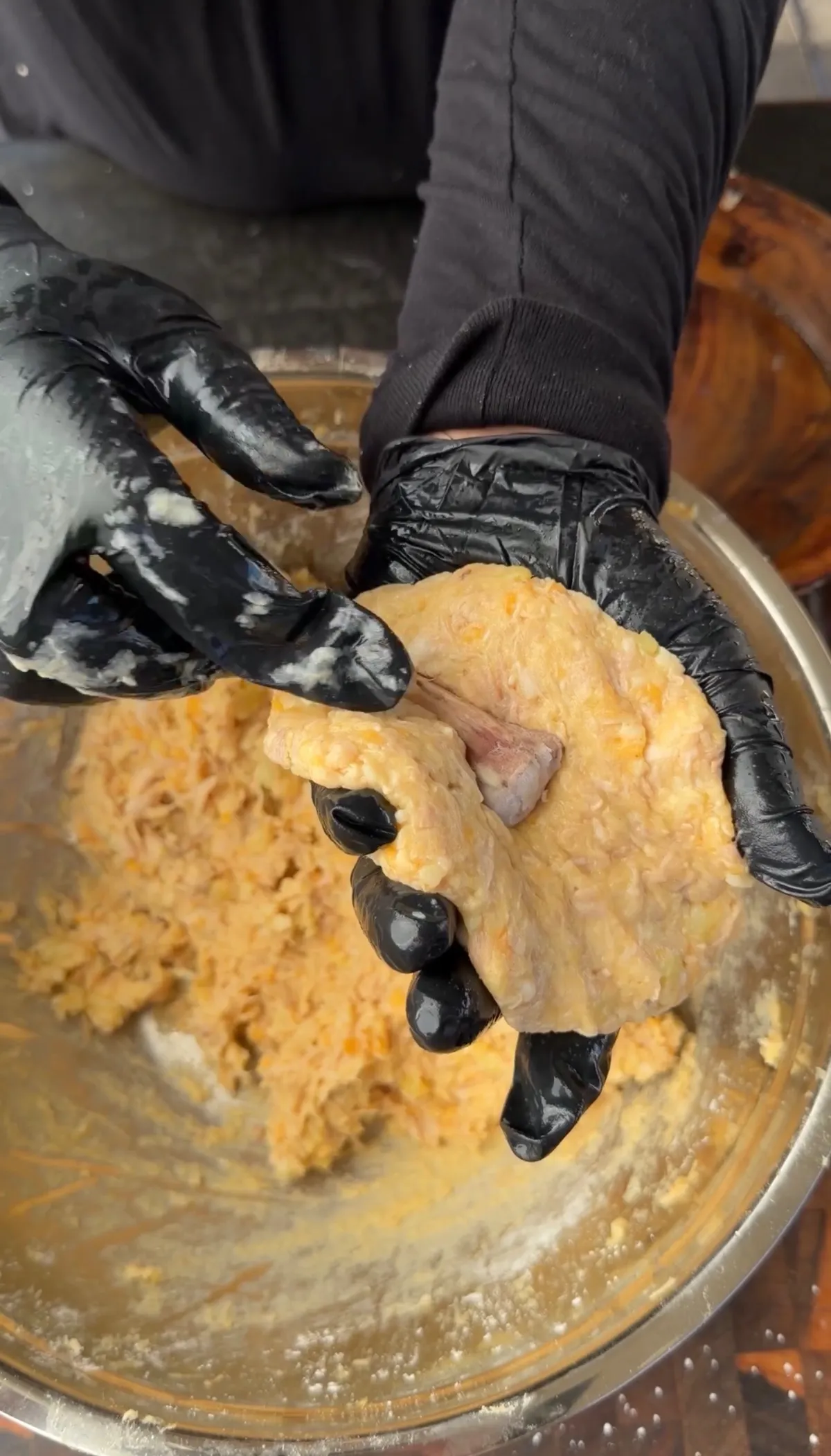 Gloved hands shaping shredded chicken and mashed potato cheese mixture into a drumstick bomb around a cleaned bone in a metal bowl