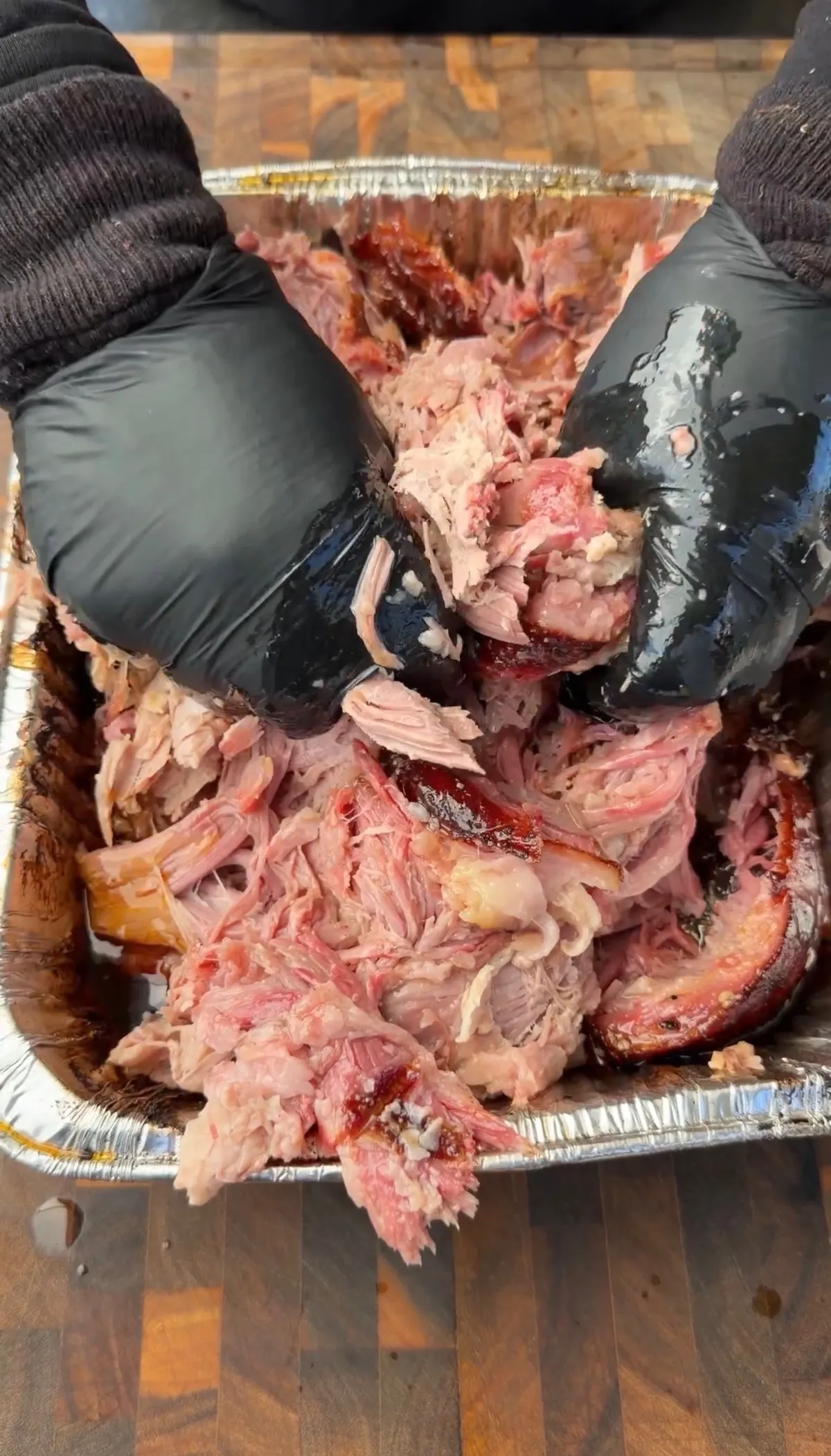 Hands in black gloves shredding smoky pulled pork shoulder in a foil tray on a wooden cutting board