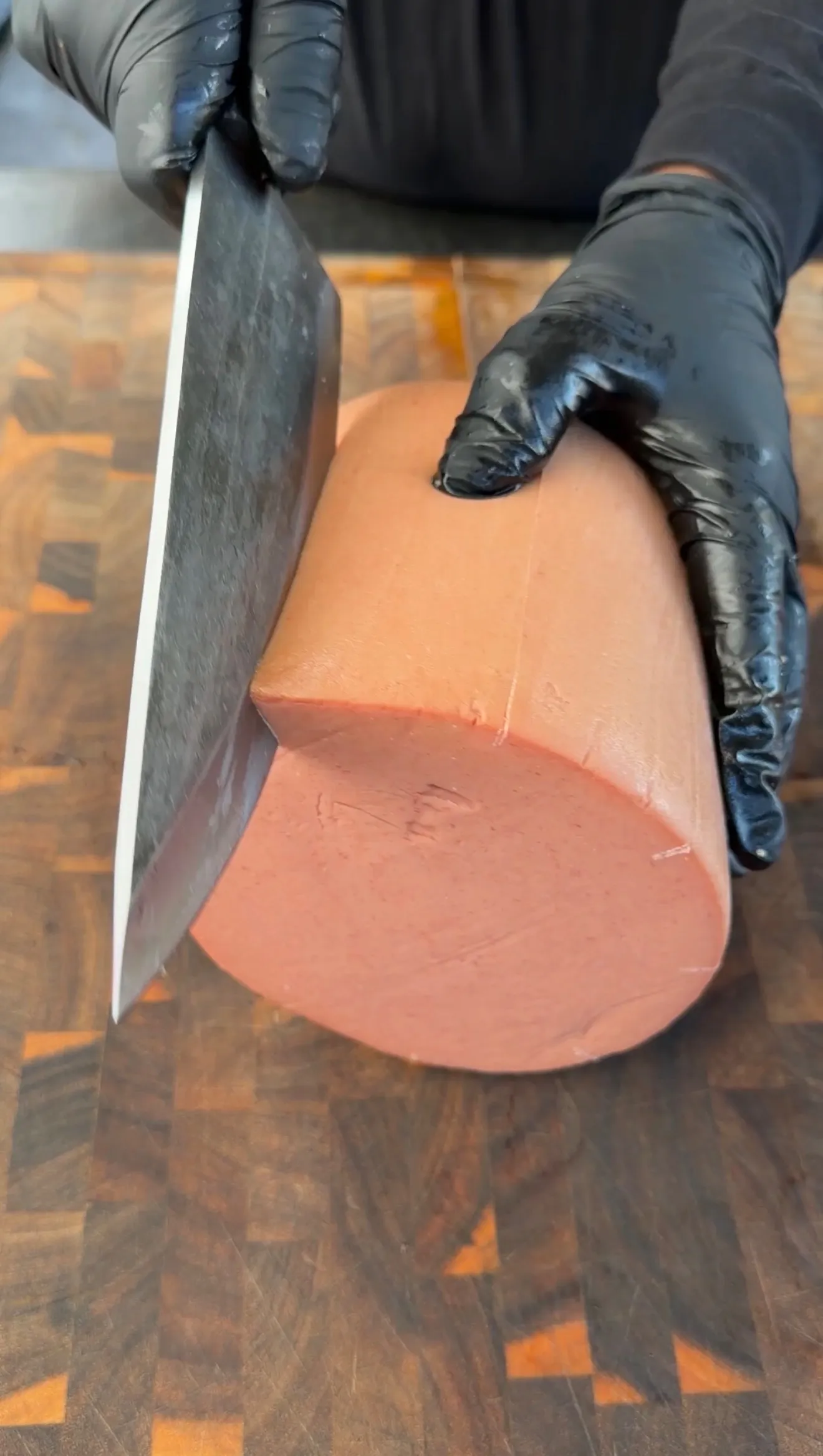 Gloved hands slicing a large bologna chub with a cleaver on a wooden end-grain cutting board