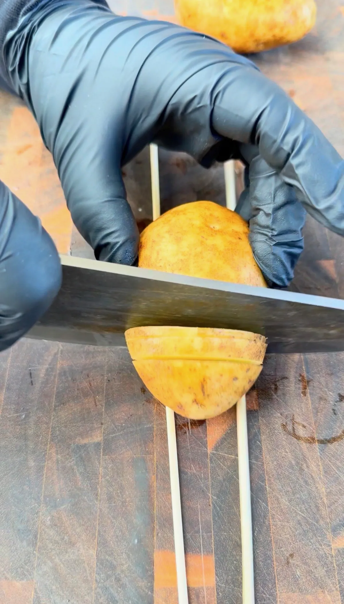 Gloved hands slicing a potato using chopstick guides on a wooden board for chicken and potato skewers