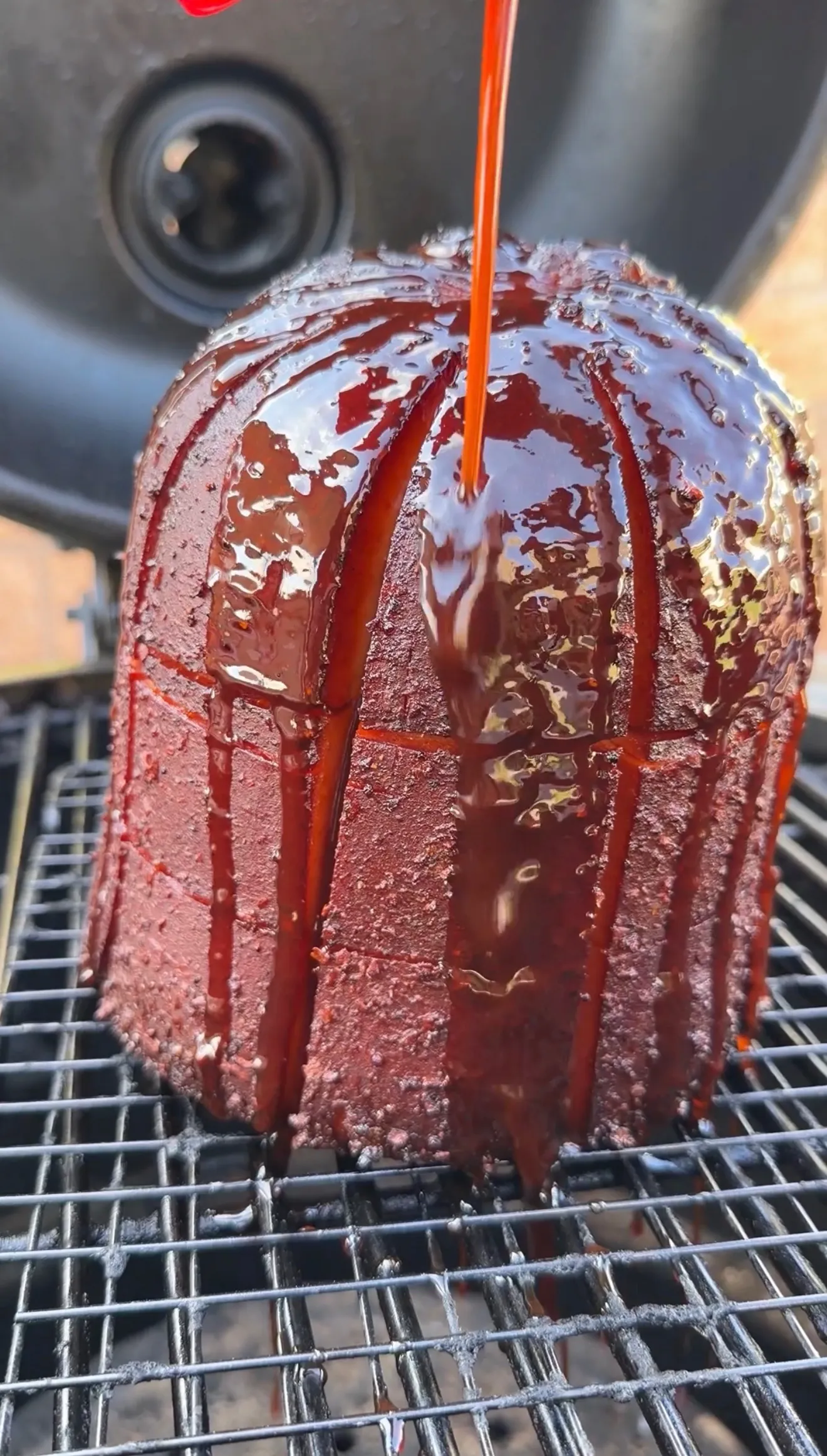 Whole smoked bologna chub with crosshatch cuts being glazed with BBQ sauce on grill grates