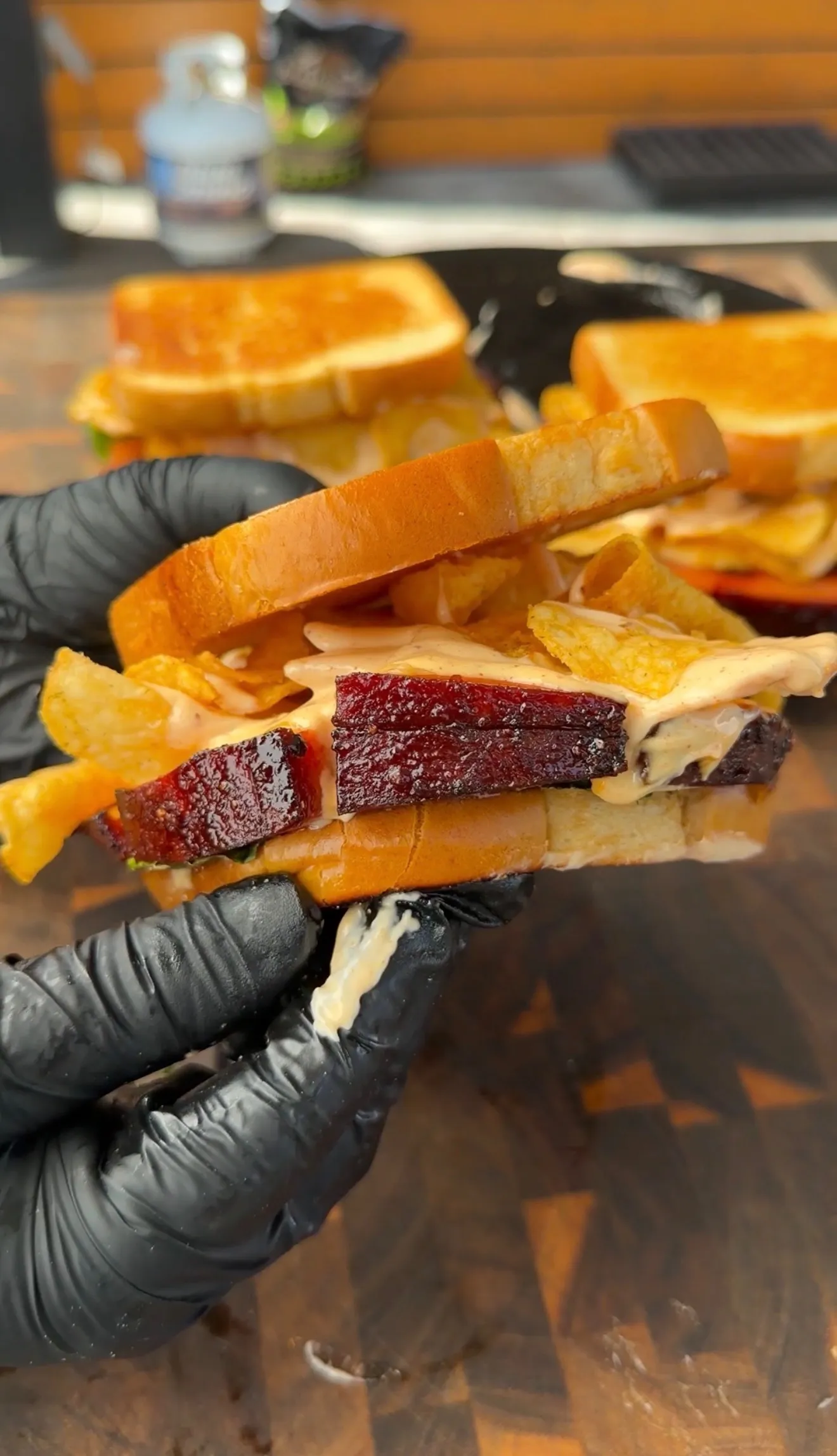 Hands in black gloves holding a thick smoked and fried bologna sandwich with chips and sauce on toasted bread